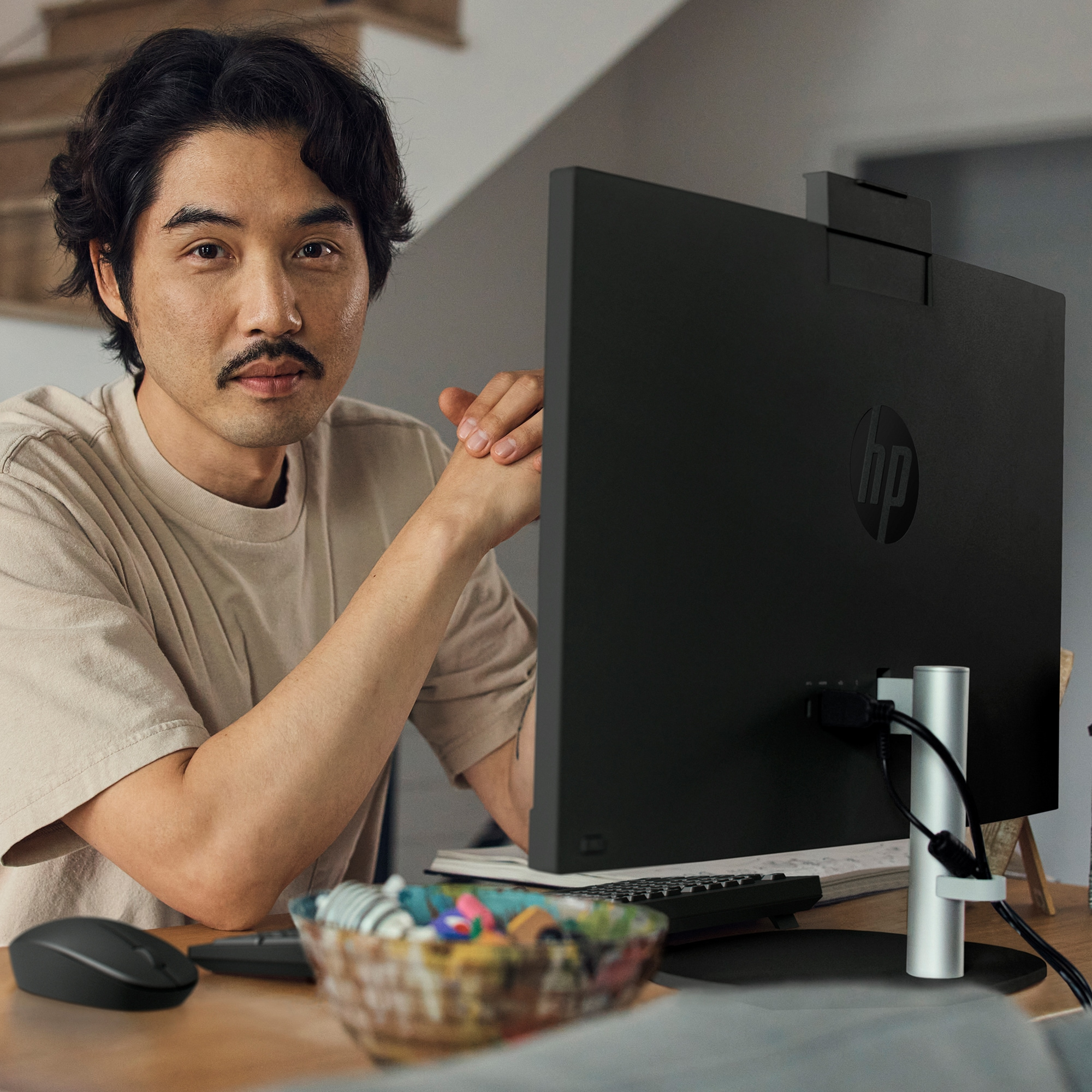 A man sits at a desk, looking at a monitor. An HP all-in-one is behind him. There is a mouse and keyboard.