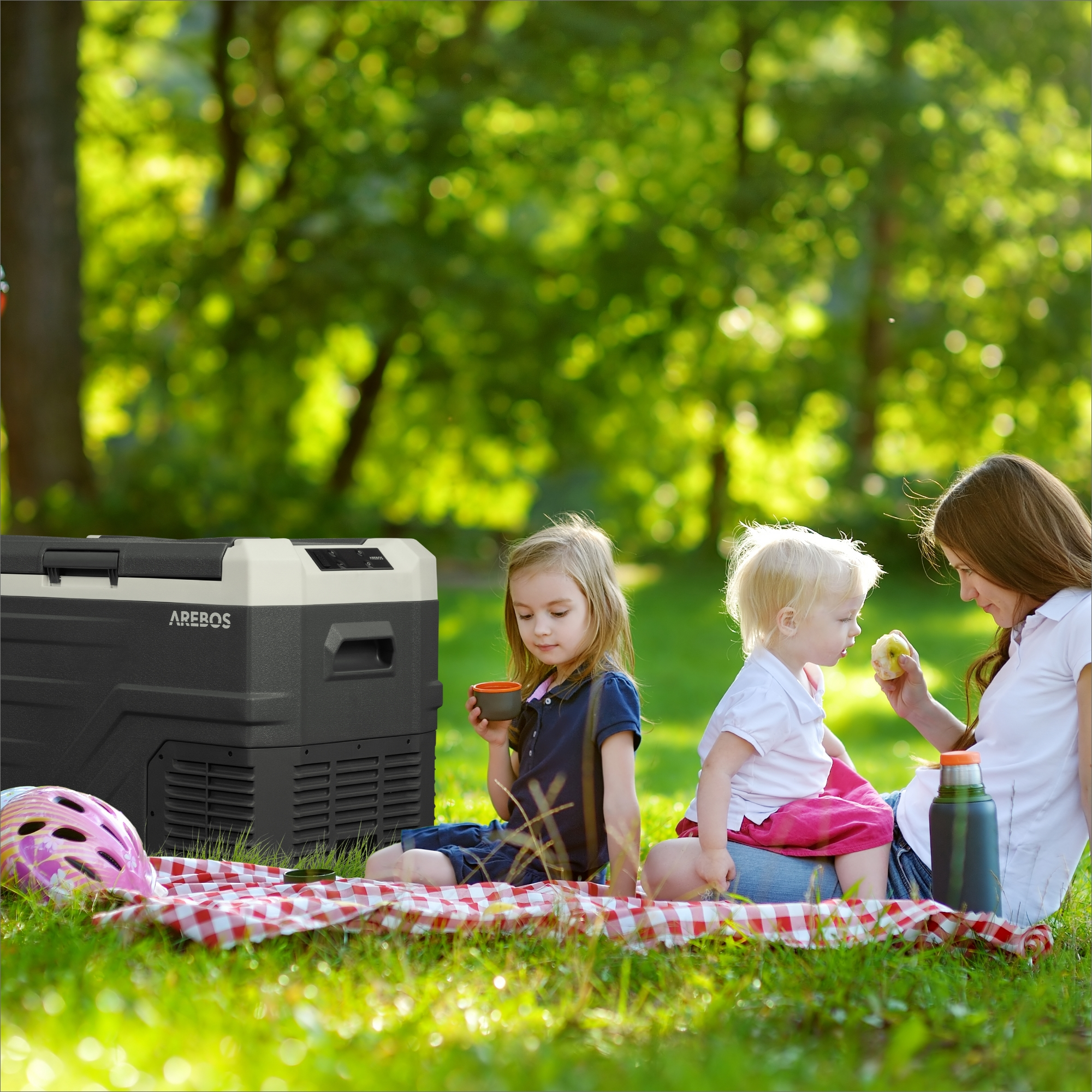 Zwei Kinder und ein Erwachsener auf einer Picknickdecke im Park mit Kühler und Picknickkorb.