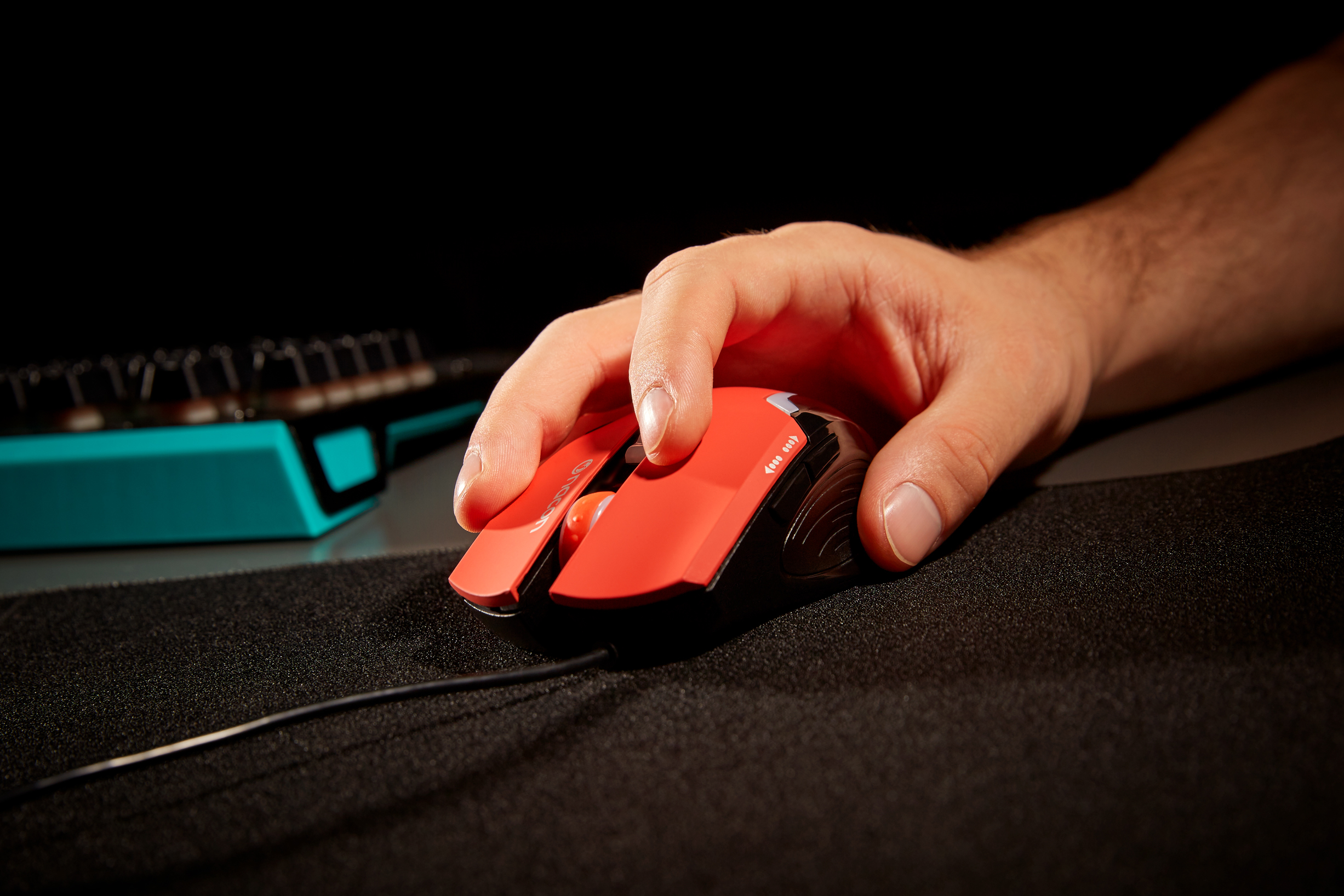 Ręka używająca czerwono-czarnej myszy do gier na czarnej podkładce, z klawiaturą w tle.