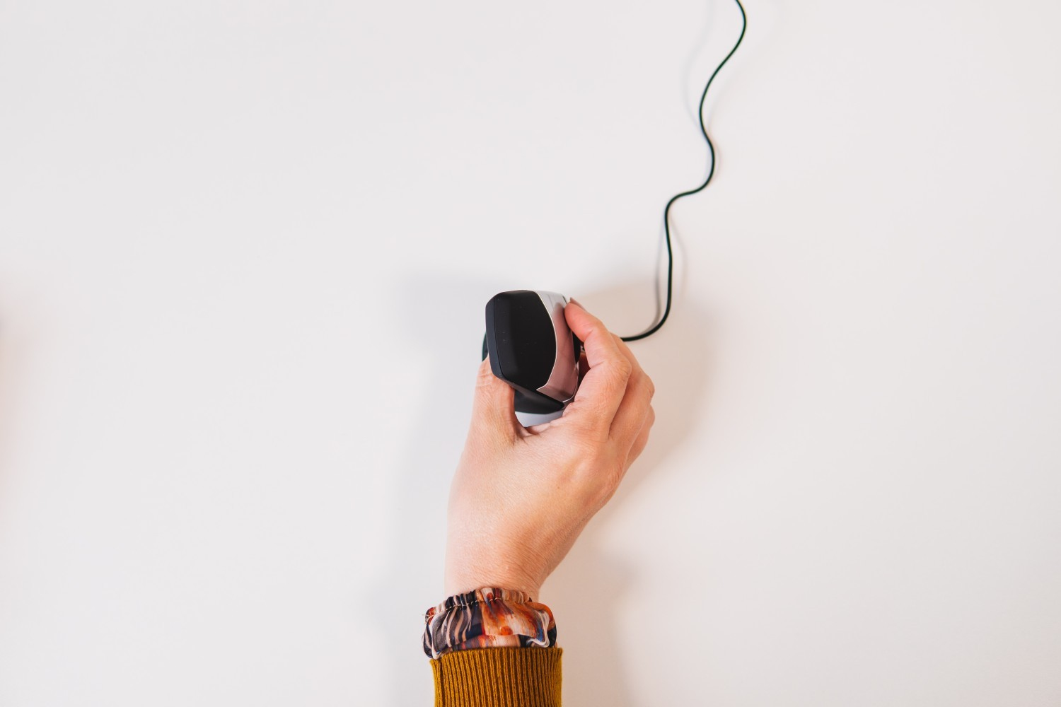 A hand holding a black and silver mouse with a black cable on a white surface.