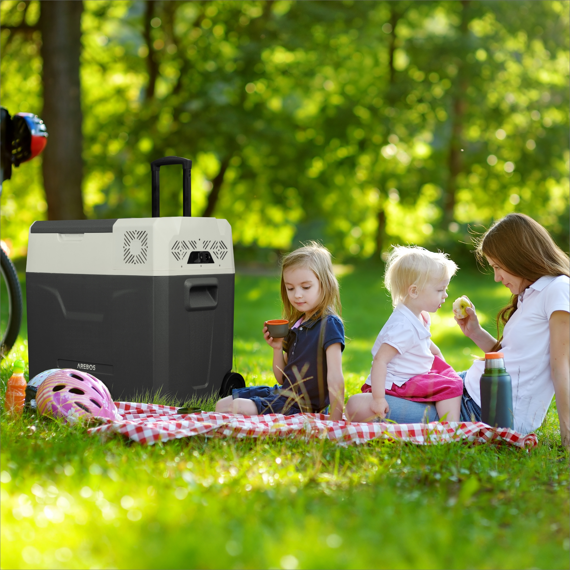 Eine Mutter und zwei Kinder picknicken in der Nähe einer Kühlbox in einem Park.