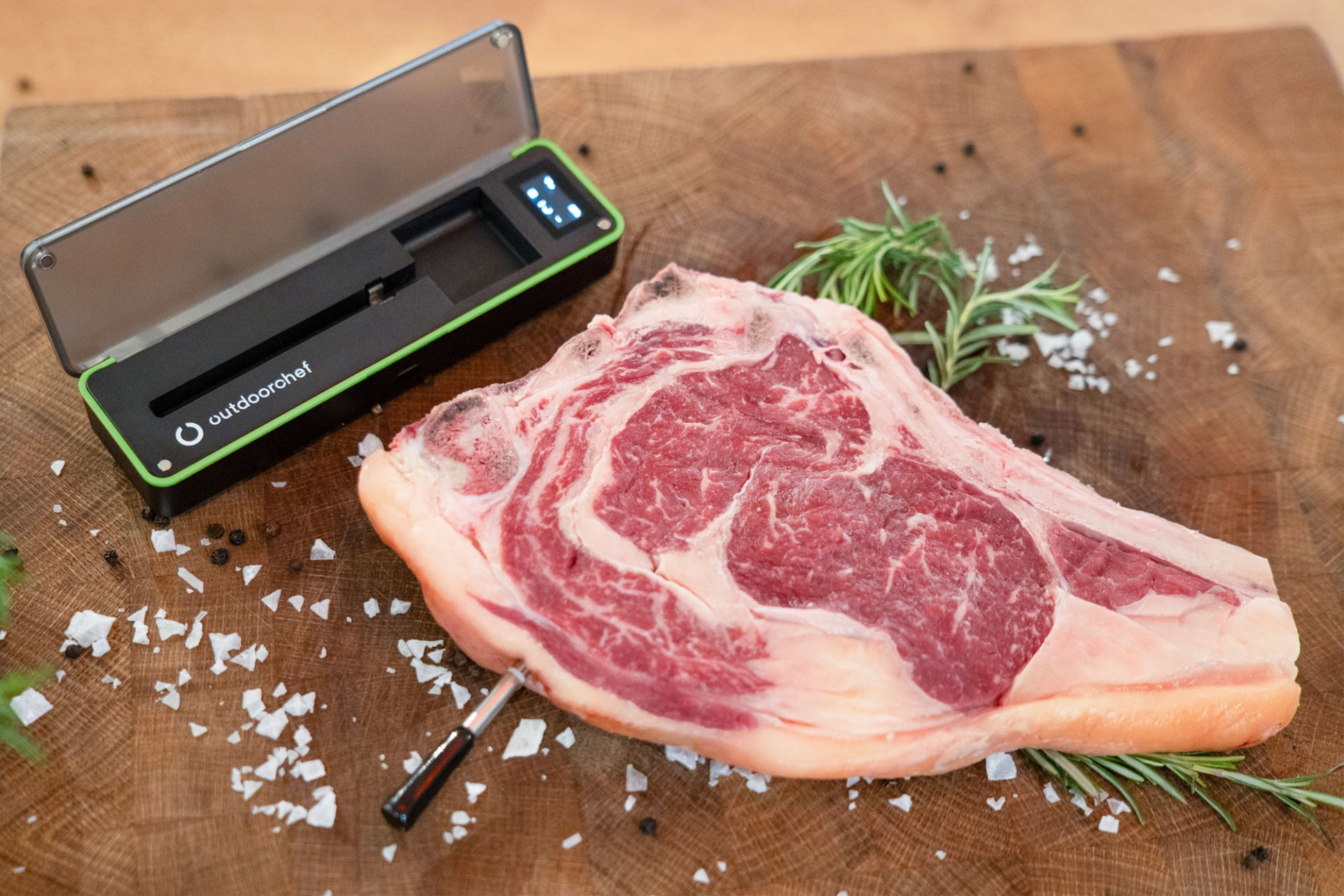 Ein rohes Steak, ein digitales Fleischthermometer und Rosmarin auf einem Holzschneidebrett.