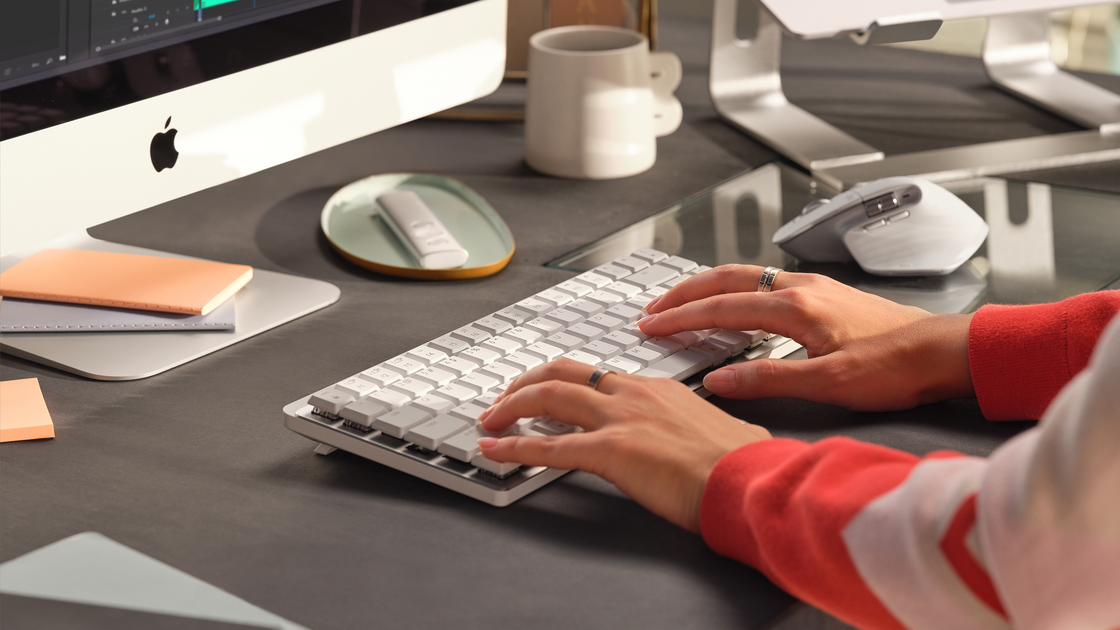 Manos escribiendo en un teclado blanco cerca de un ordenador y otros objetos.