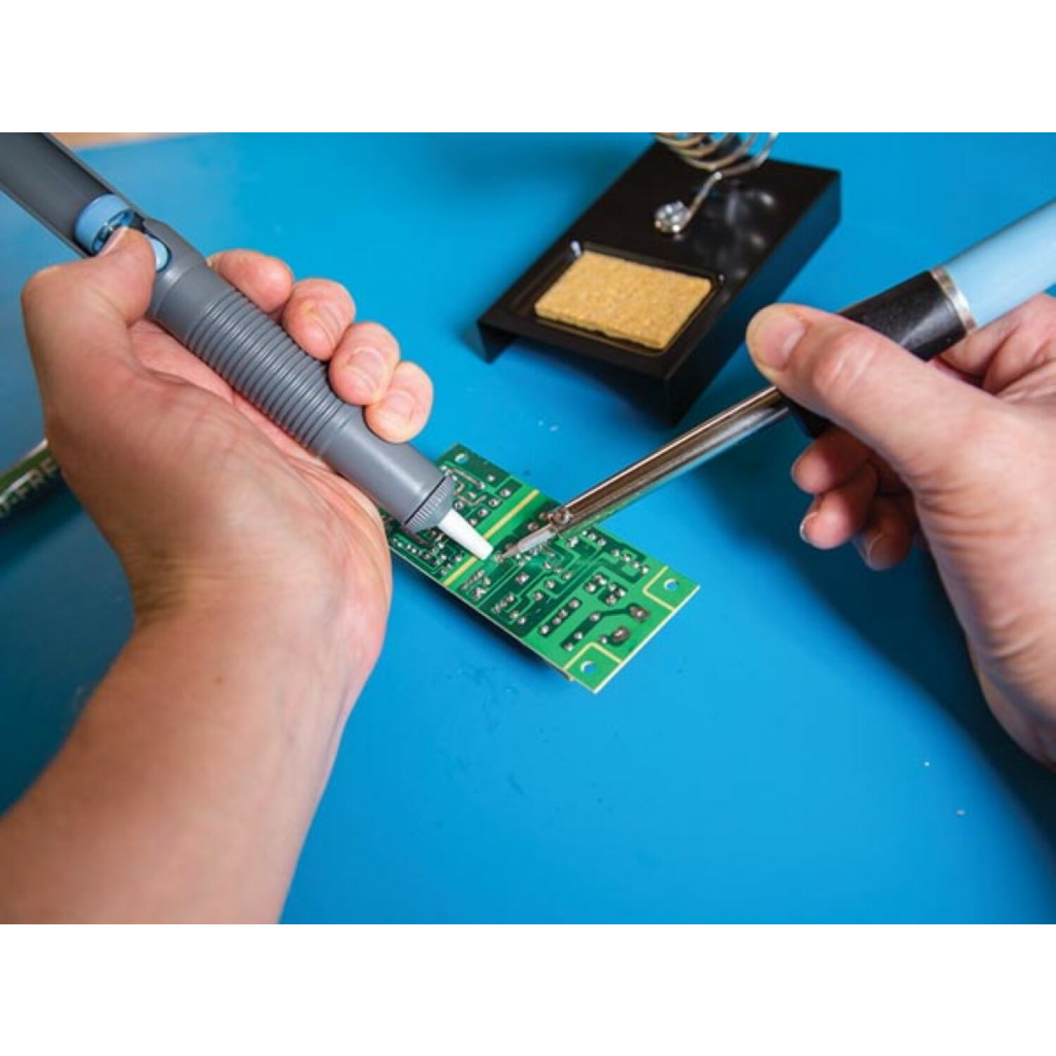 Mani che saldano su un circuito stampato. Saldatore e pompa dissaldante su superficie blu.
