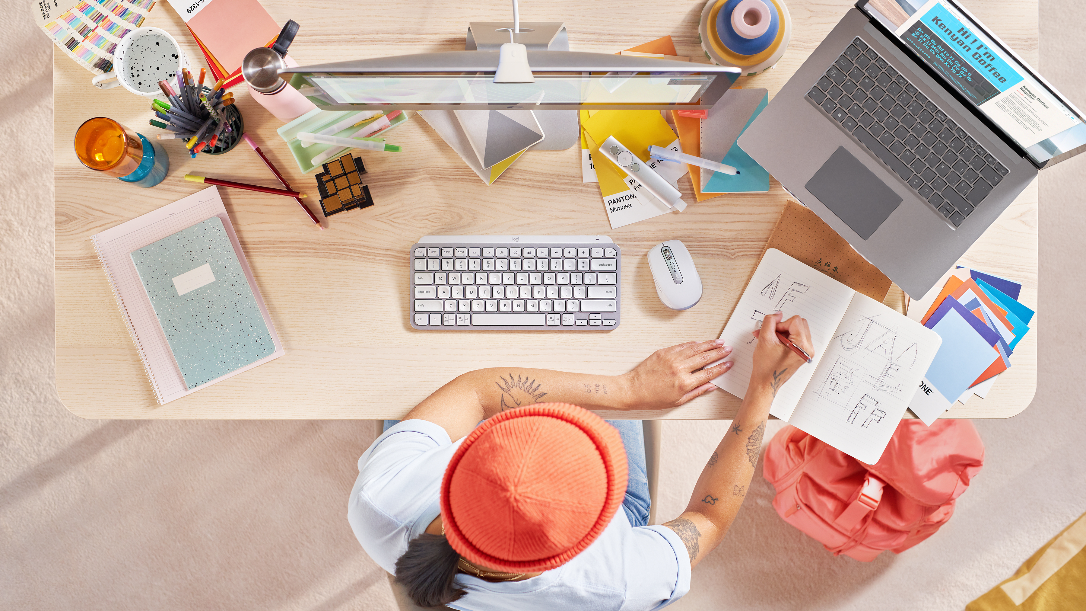 Widok z góry na biurko z laptopem, klawiaturą, notatnikiem, długopisami i osobą rysującą.