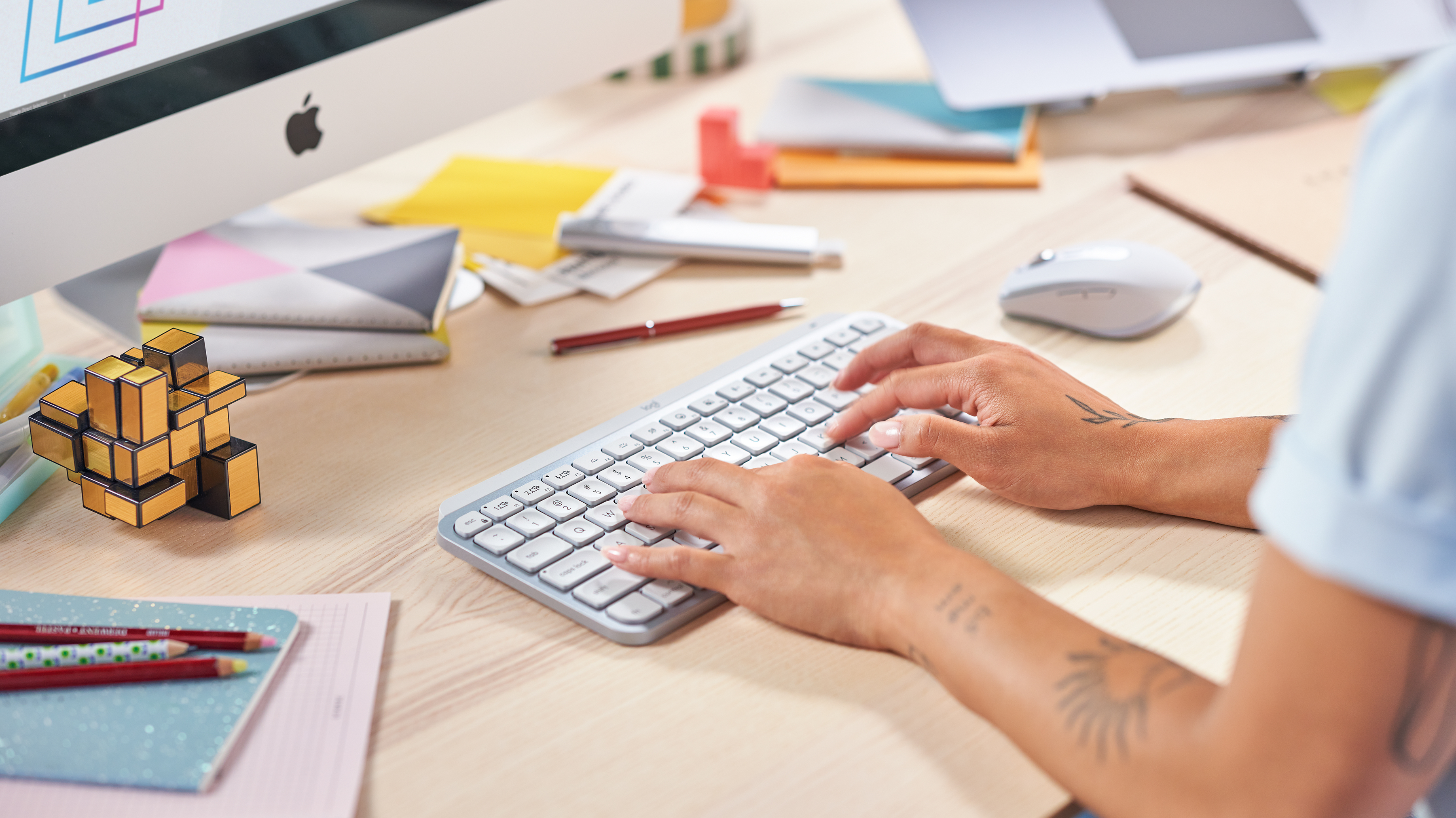 Ręce piszące na białej klawiaturze, z ekranem komputera i innymi materiałami biurowymi na drewnianym biurku.