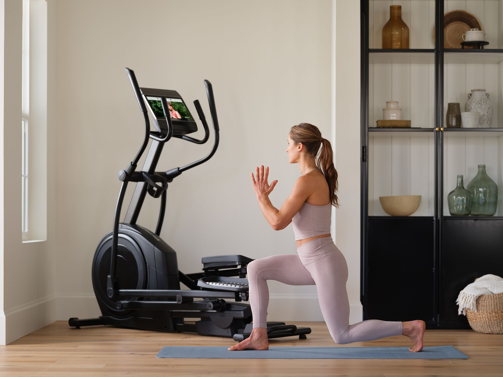 Frau in Sportkleidung macht einen Ausfallschritt neben einem Crosstrainer. Heller, moderner Raum.