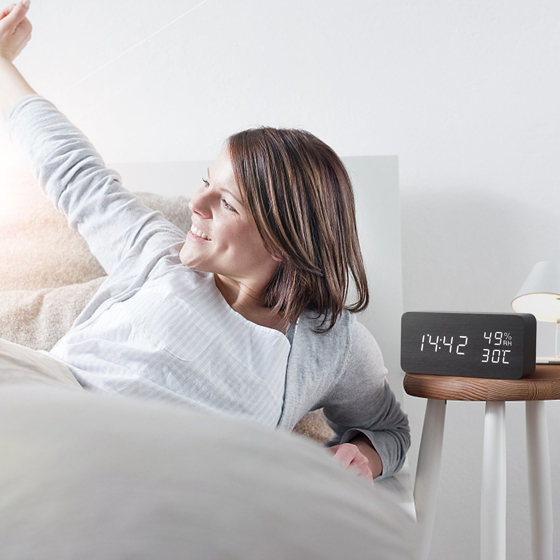 Frau im Bett greift nach einer digitalen Holzuhr mit Zeit- und Datenanzeige.