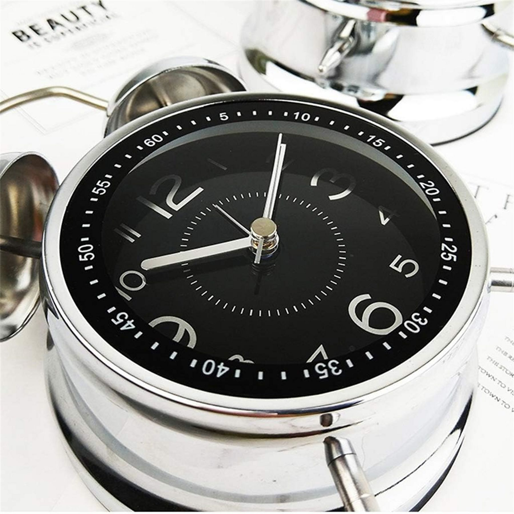 Ein silberner Wecker mit schwarzem Zifferblatt. Die Uhr zeigt die Zeit an. Eine weitere Uhr ist im Hintergrund.