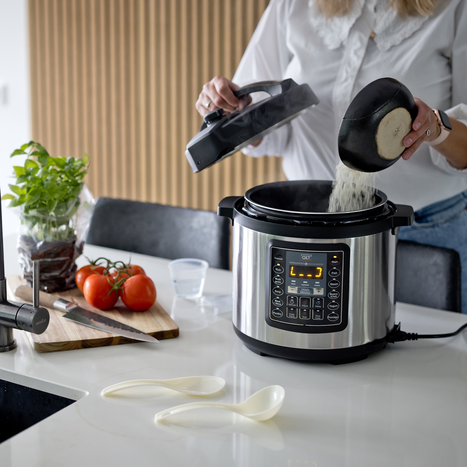 Woman pouring rice from a pot into a silver electric cooker, with tomatoes and spoons.