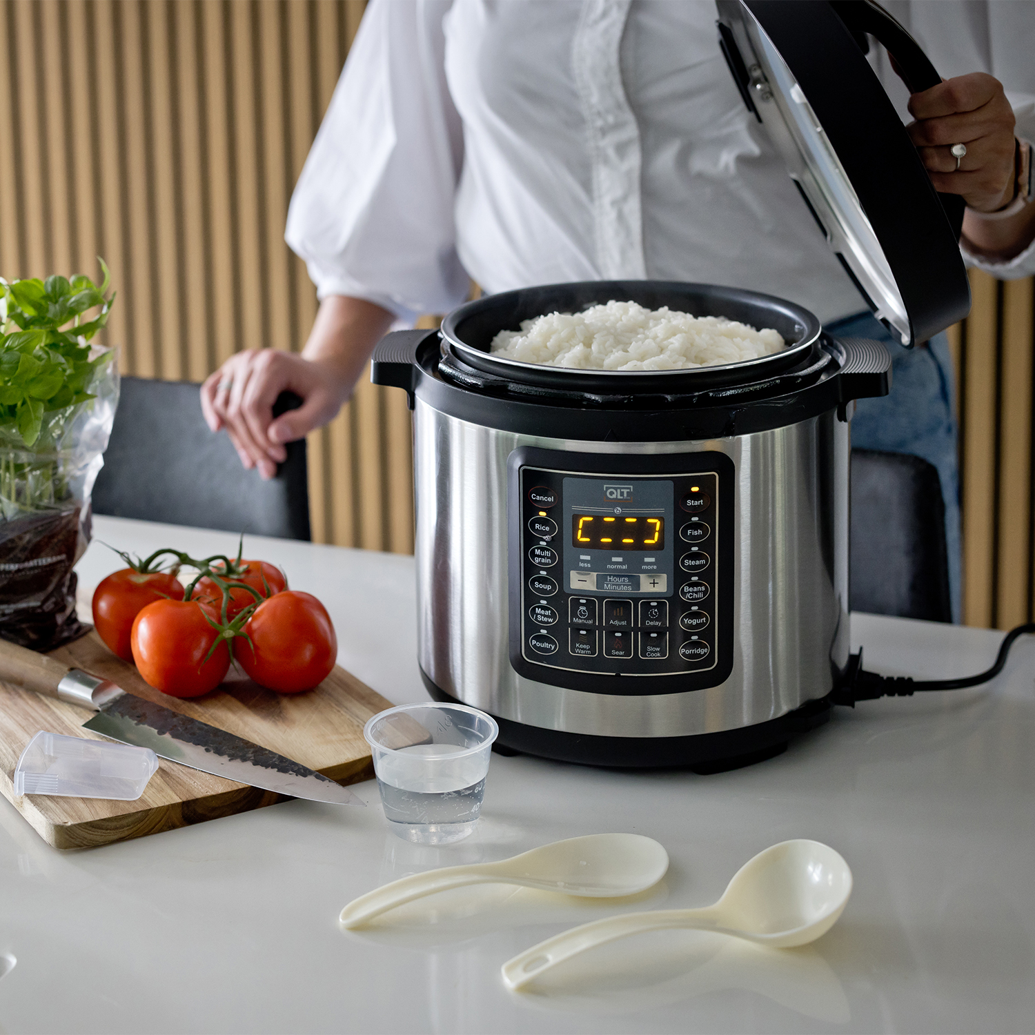 Woman near rice cooker with cooked rice. Tomatoes, knife, and spoons on the table.