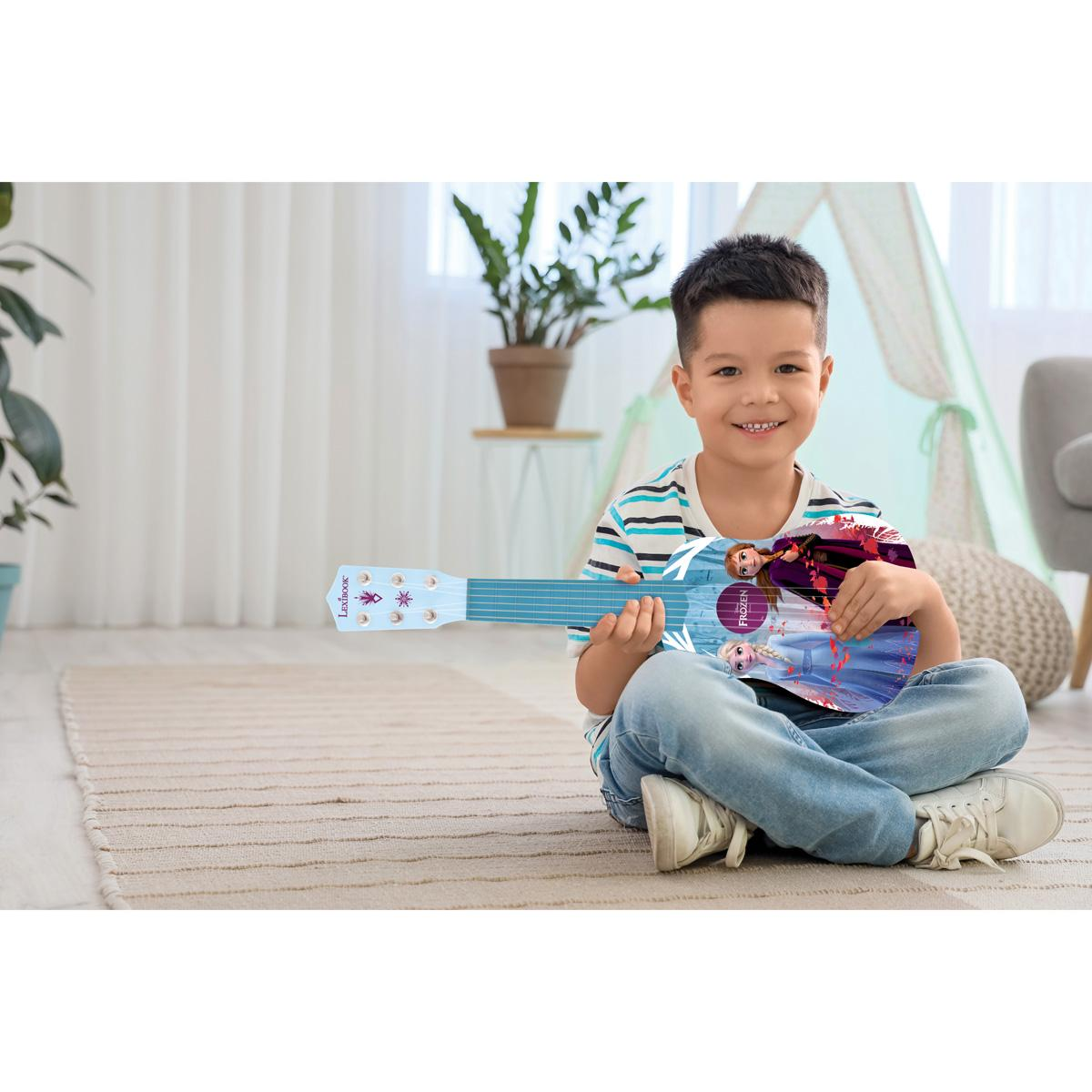 Chłopiec gra na zabawkowym gitarze z motywem Krainy Lodu. Uśmiecha się, siedząc na dywanie.