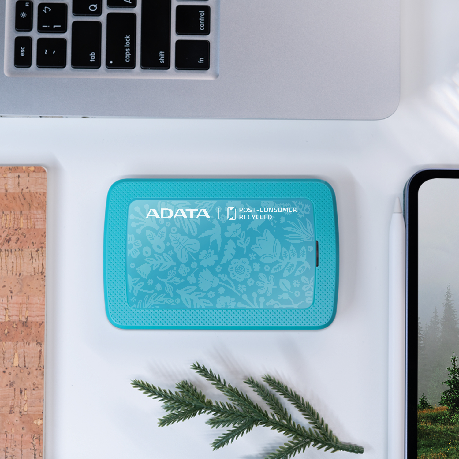Un disco duro externo ADATA turquesa sobre un escritorio blanco con un portátil y un corcho.