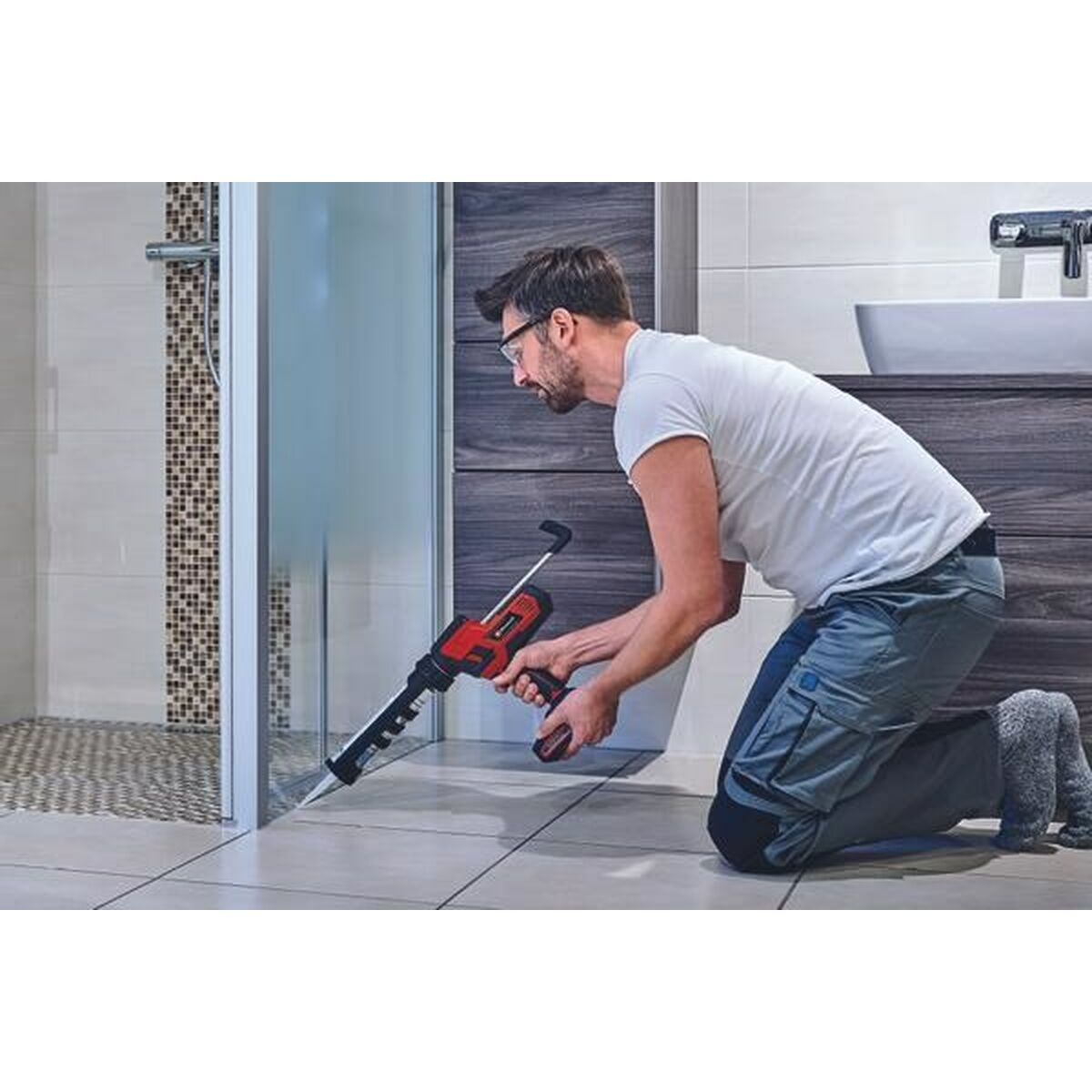 A man kneeling, using a red caulk gun, applying sealant in a bathroom.