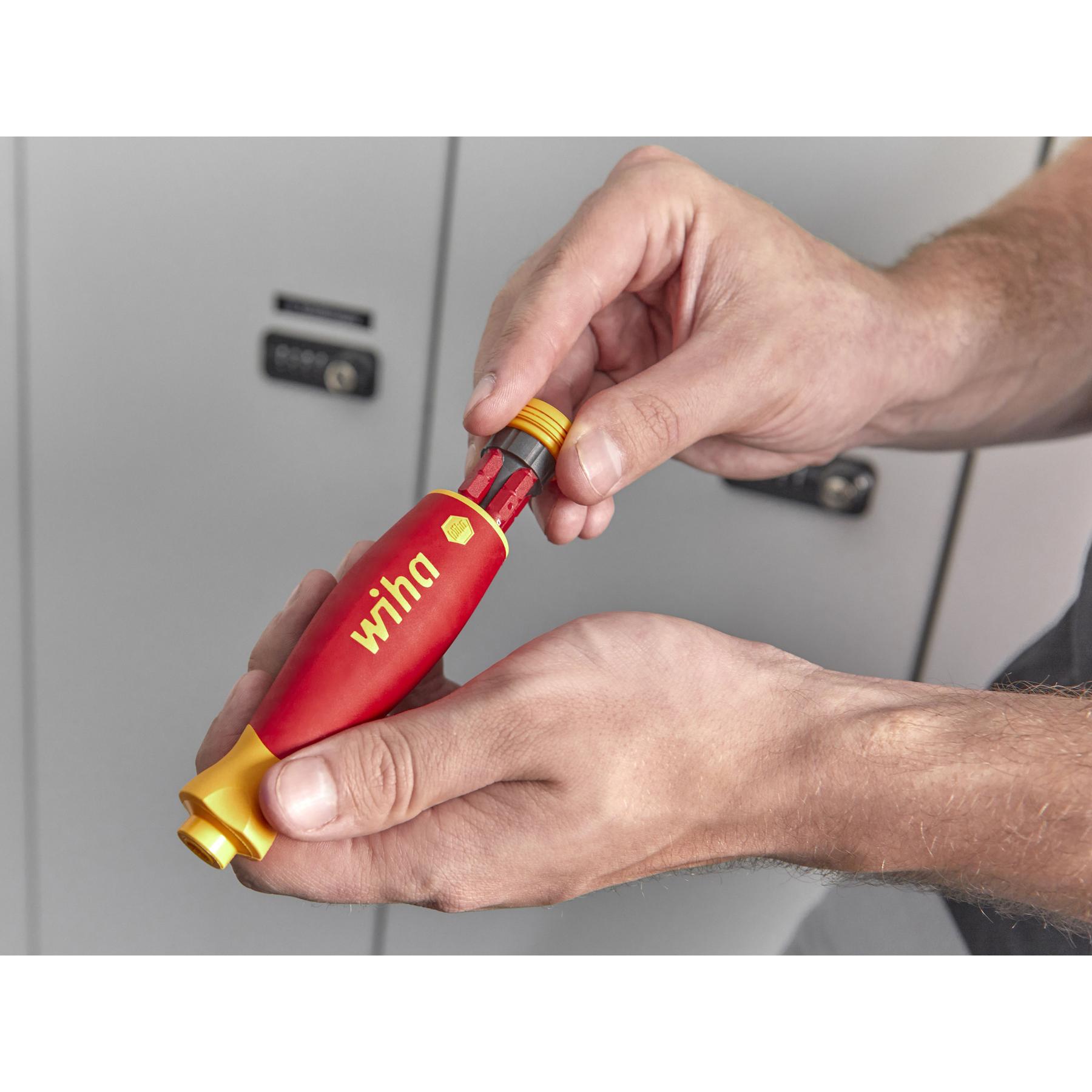 Dłonie trzymające czerwono-żółty śrubokręt Wiha, w pobliżu szarej szafki.