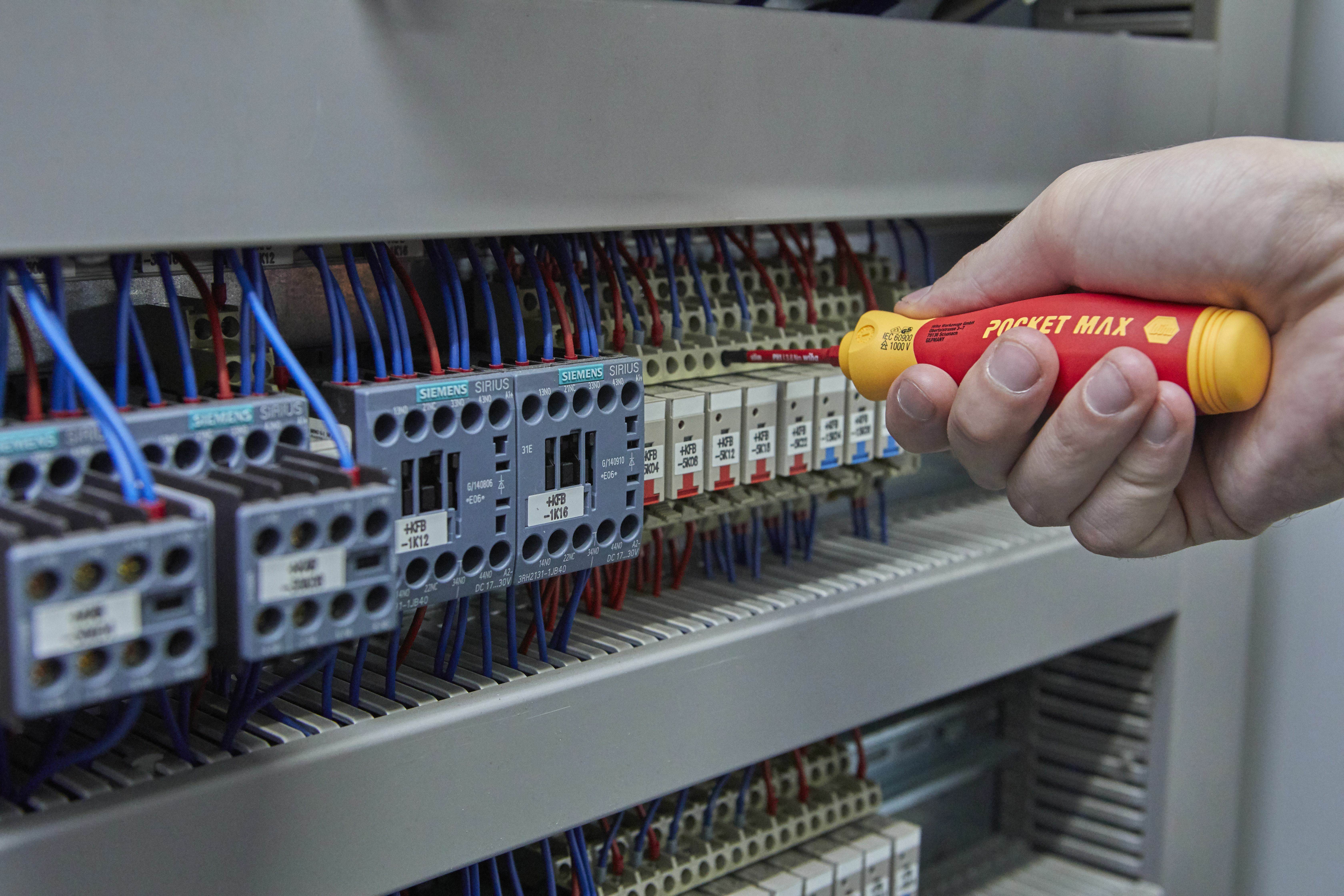 Dłoń trzymająca czerwono-żółty śrubokręt przy panelu elektrycznym z przewodami.