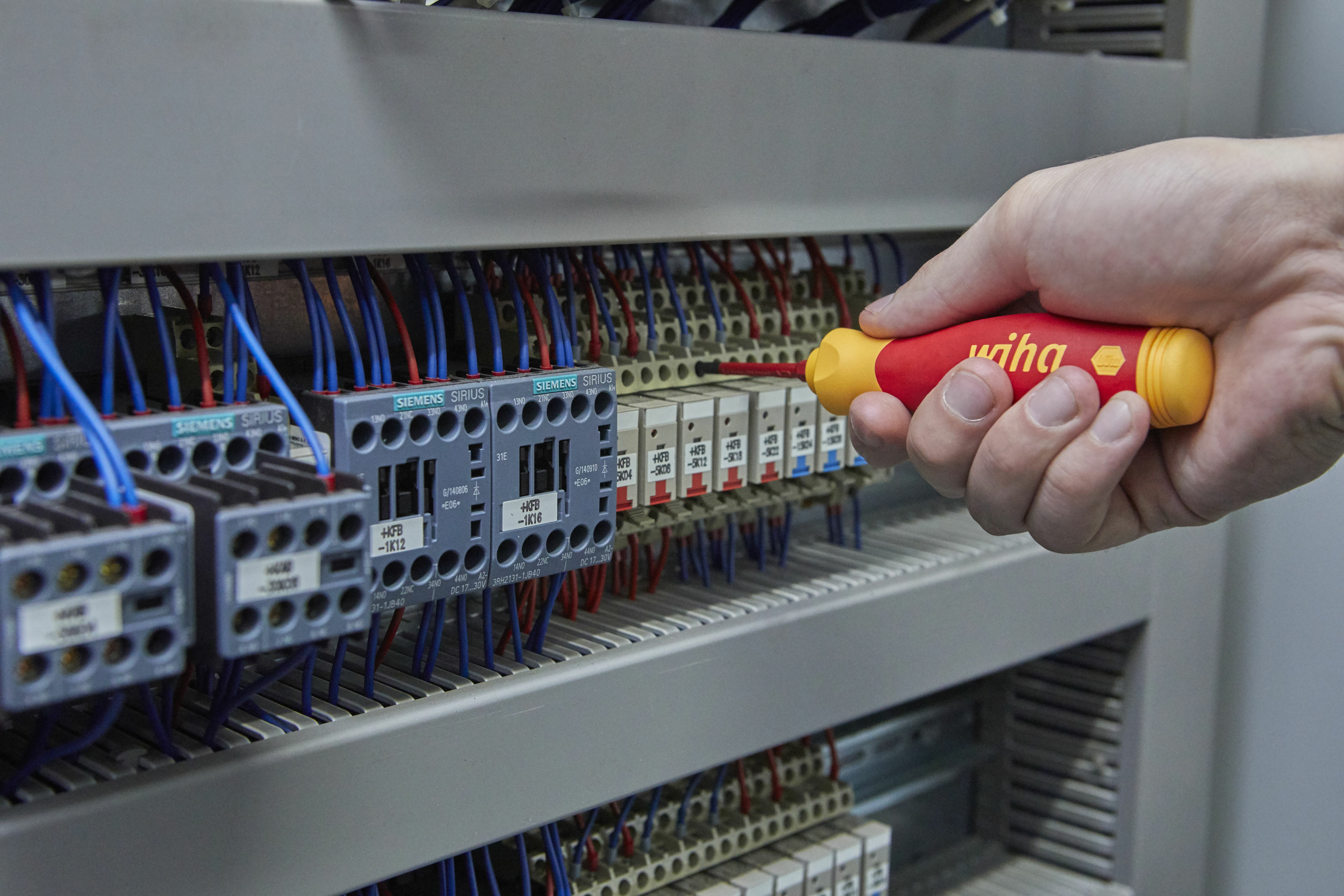 Dłoń używająca czerwono-żółtego śrubokrętu Wiha na panelu elektrycznym z przewodami.