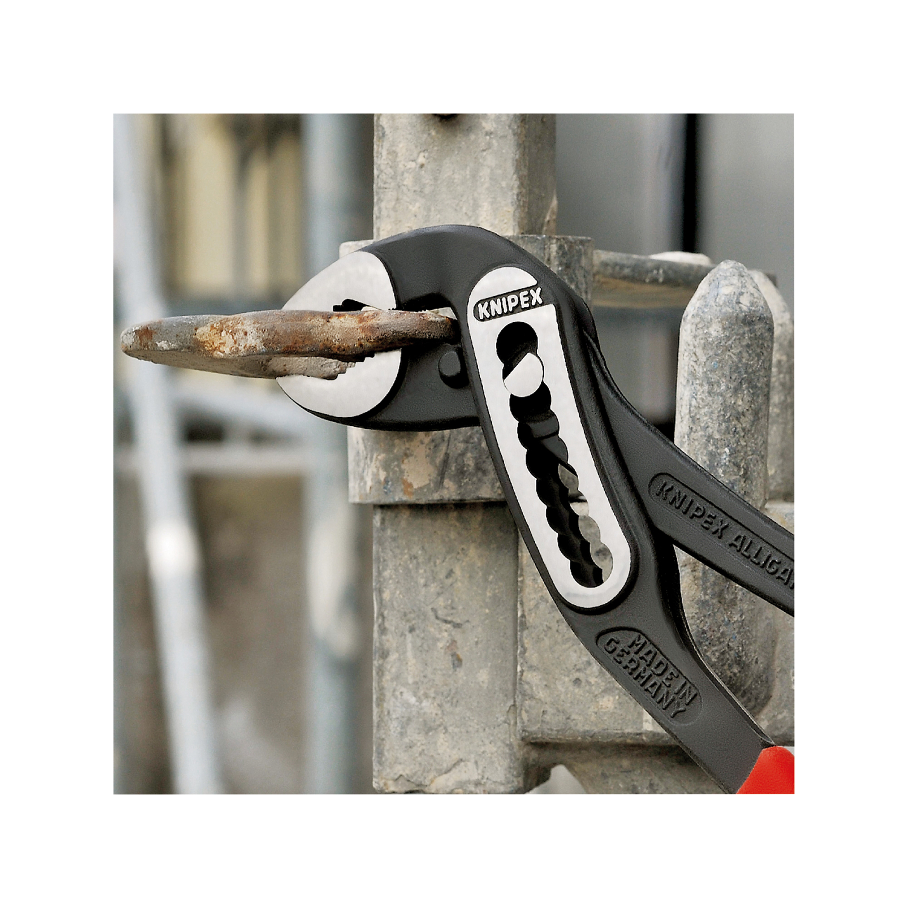 Szczypce Knipex chwytające metalowy pręt, czerwone uchwyty, na tle metalowej konstrukcji.