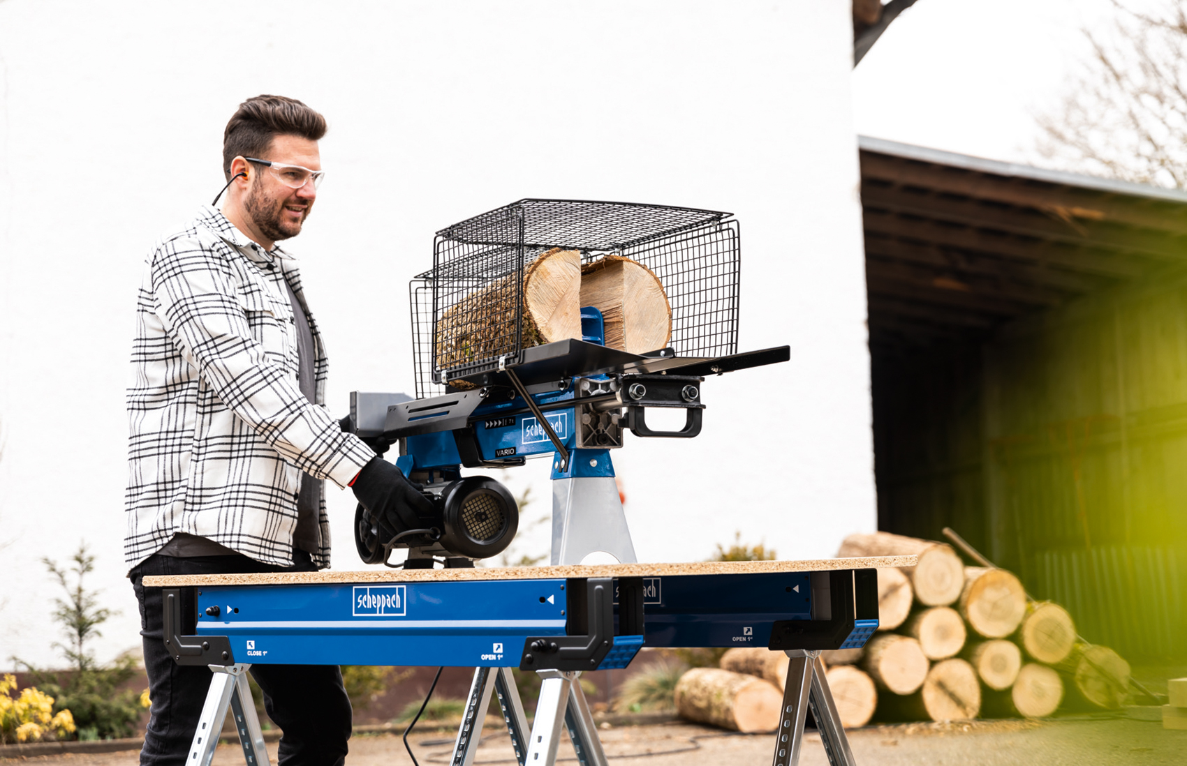 Mann benutzt eine Holzspaltmaschine mit einem Stapel Holz im Hintergrund.