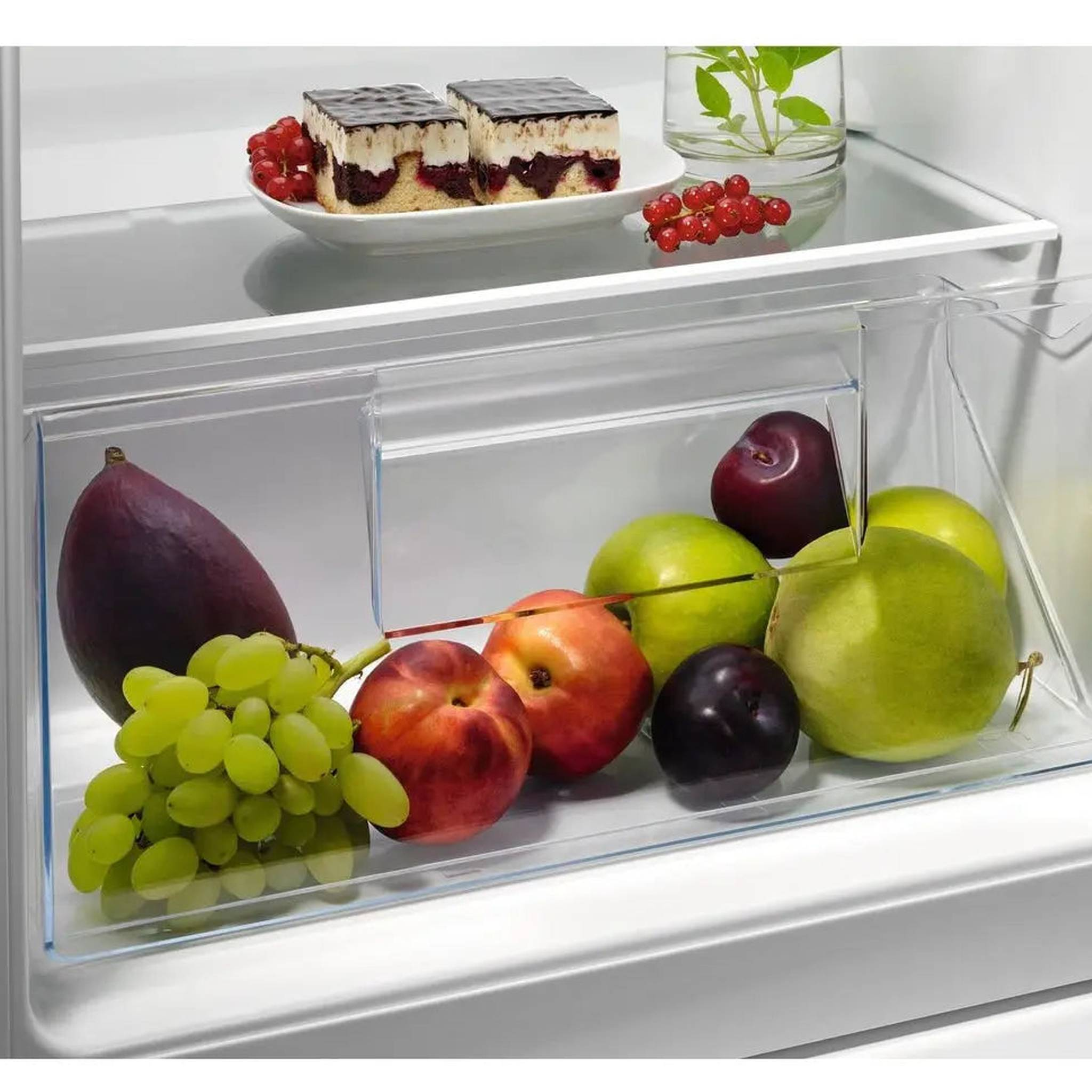 Cassetto del frigorifero con frutta e dolci, tra cui uva, prugne, mele e un dessert.