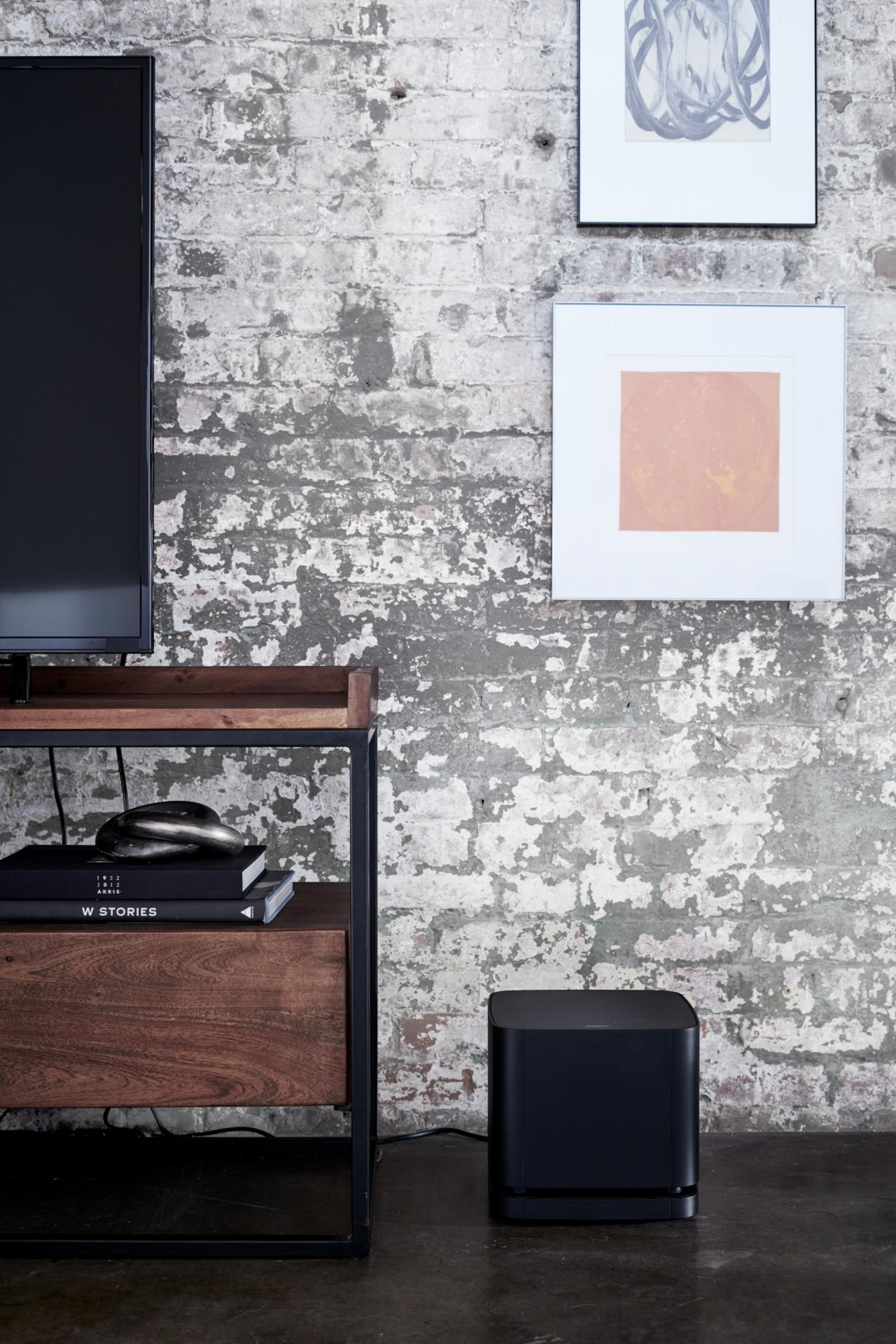 Un subwoofer Bose negro se asienta en un suelo oscuro contra una pared de ladrillos blancos desgastados.