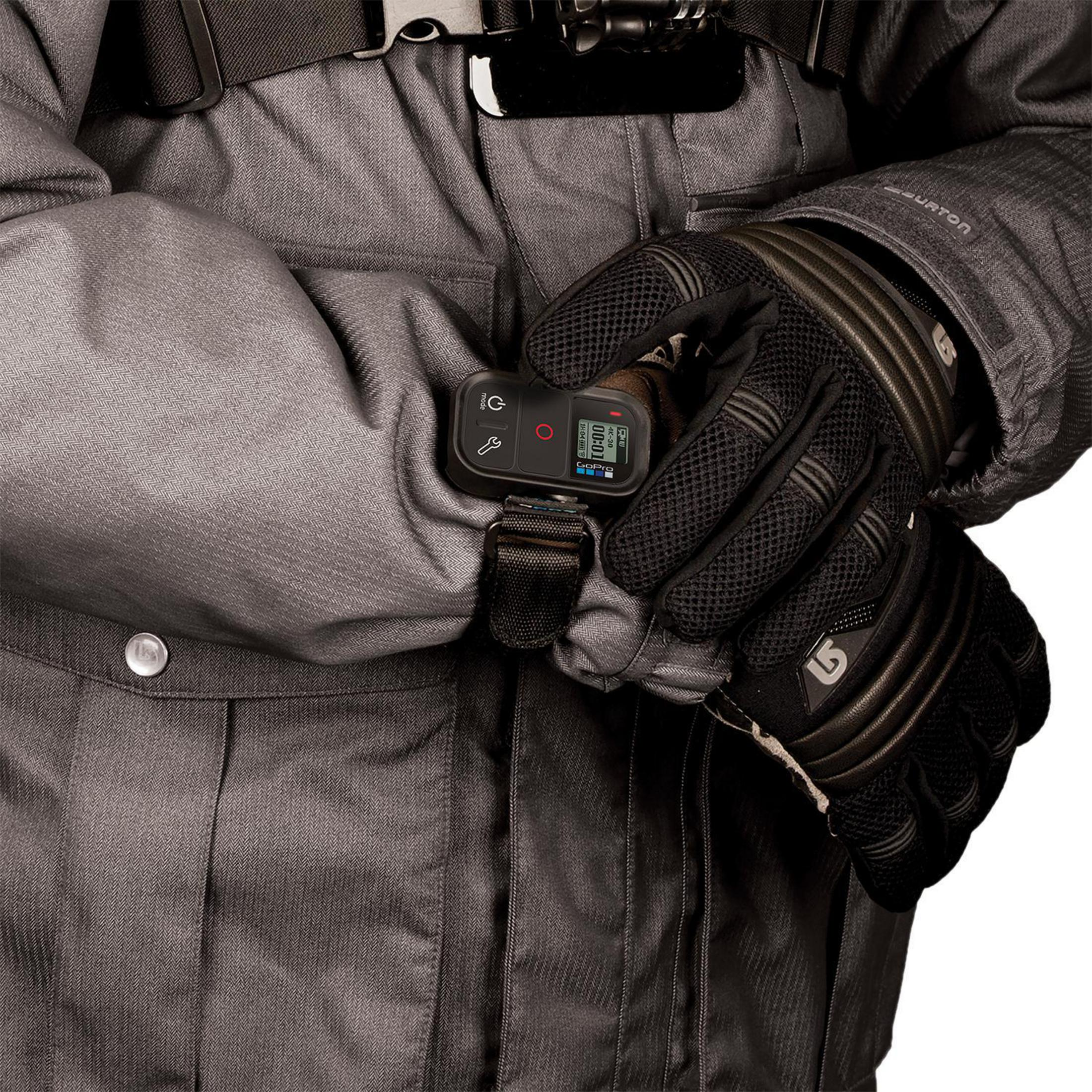 Osoba w szarej kurtce i czarnych rękawicach, trzymająca czarny pilot GoPro z ekranem.