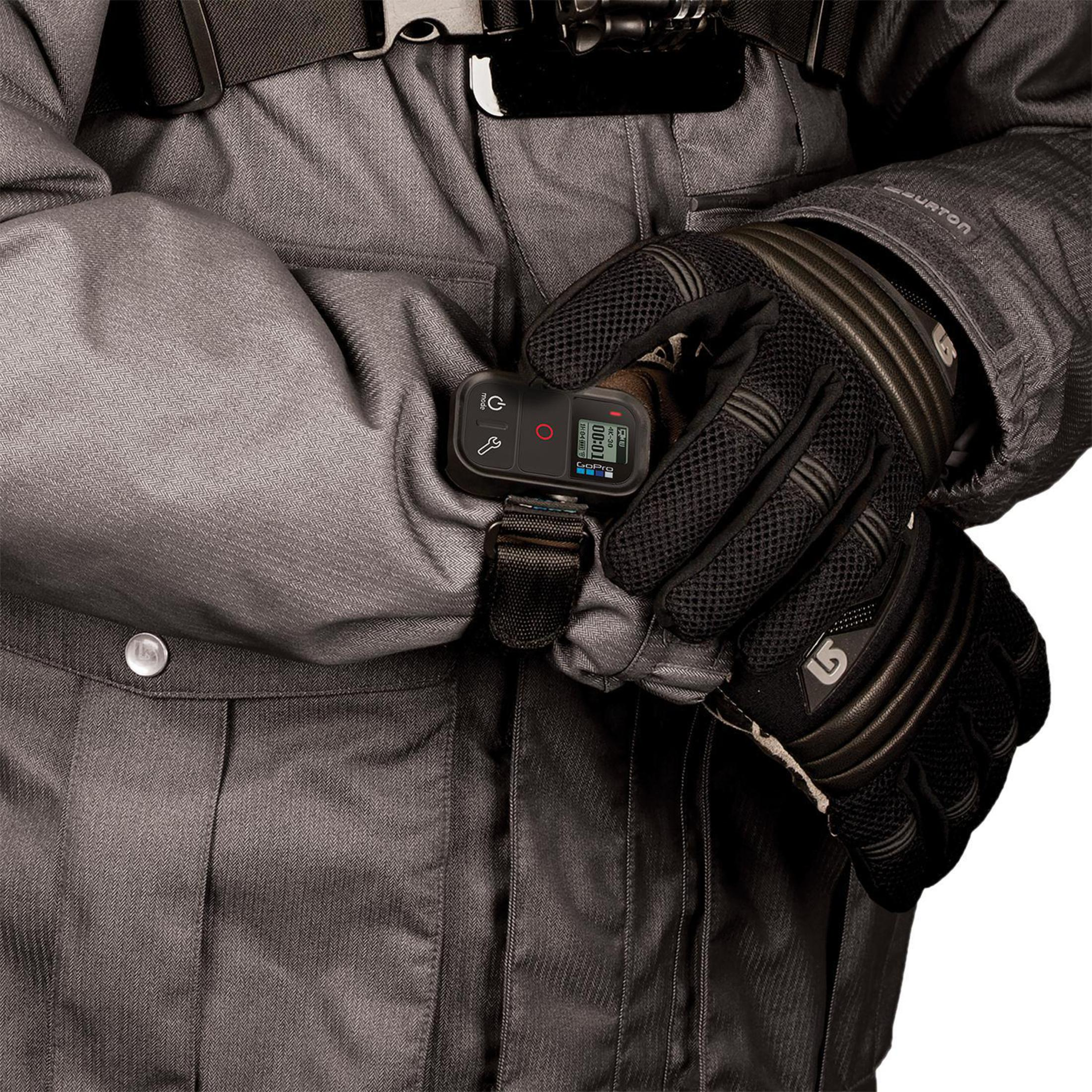 Osoba ubrana w szare ubrania zimowe i czarne rękawice, trzymająca czarny pilot do kamery sportowej.