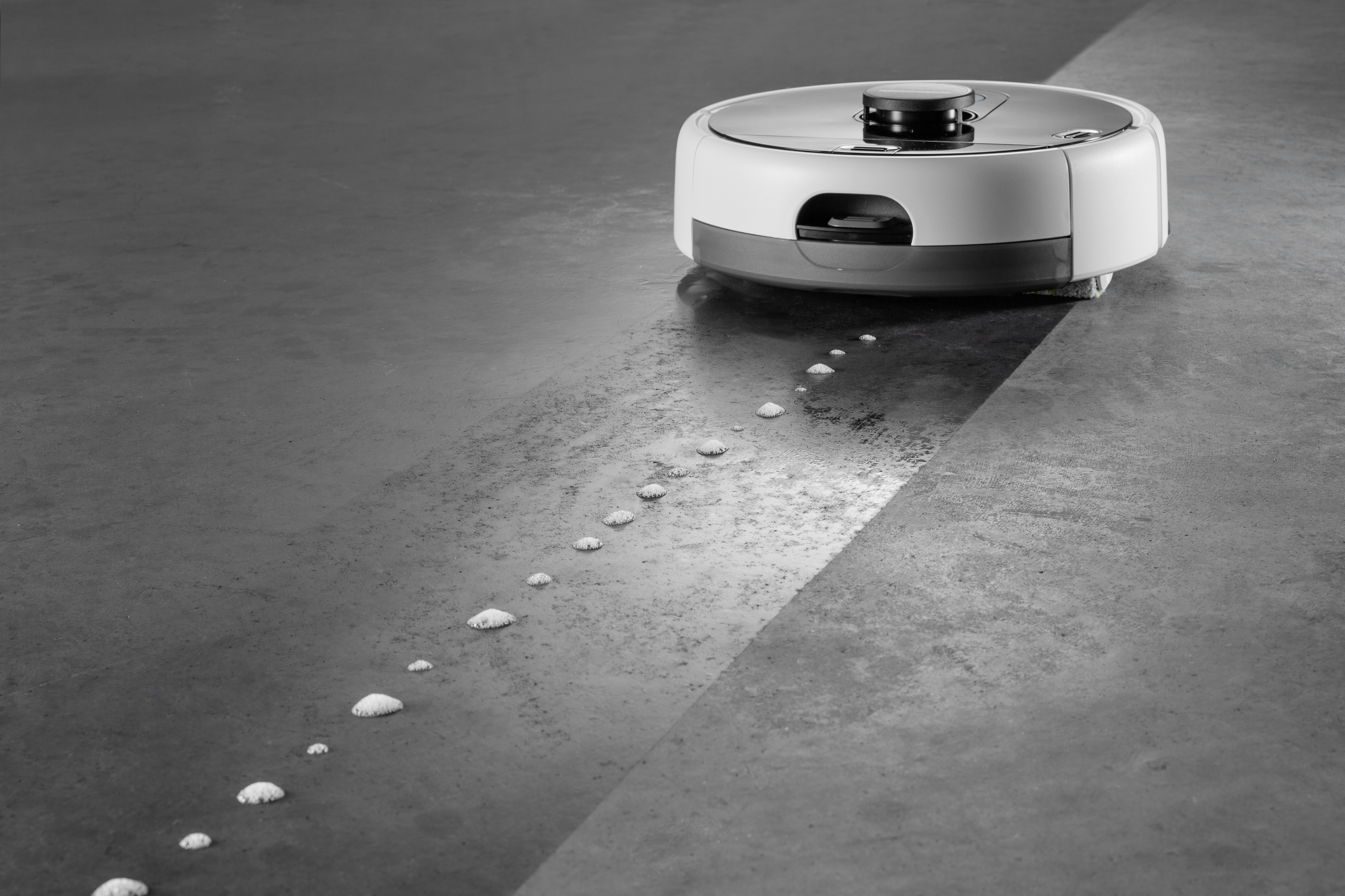 Biały odkurzacz robot na szarej podłodze, pozostawiający ślad kropelek wody.