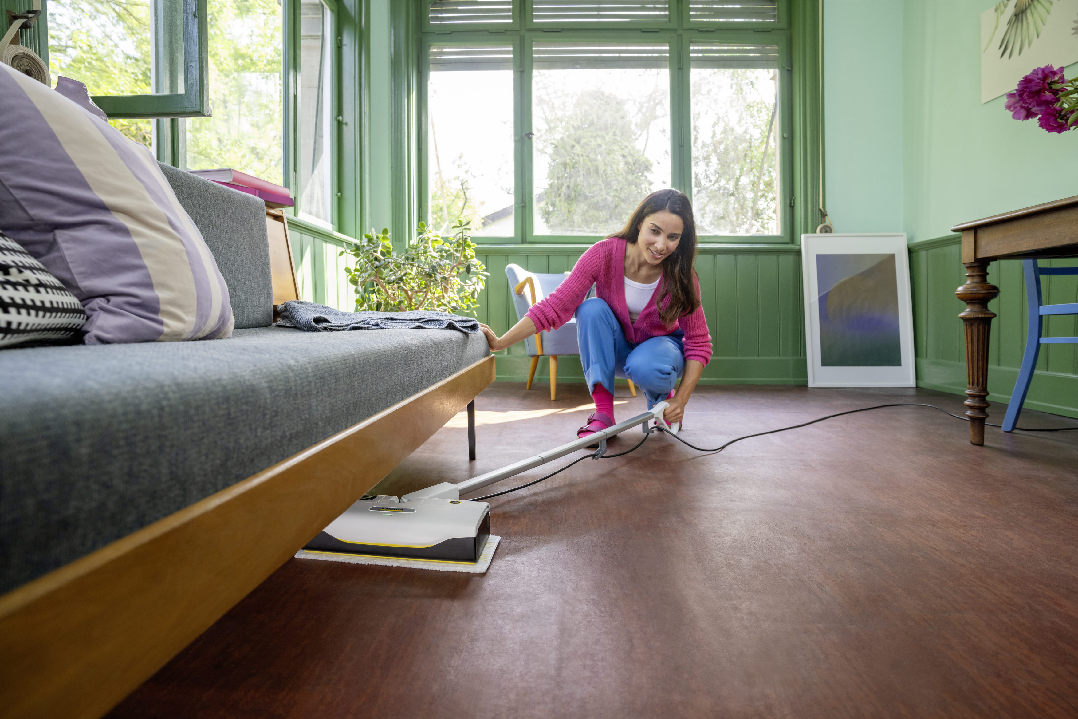 Kobieta czyszcząca drewnianą podłogę za pomocą środka do czyszczenia podłóg. W pokoju są okna, sofa i kilka dzieł sztuki.