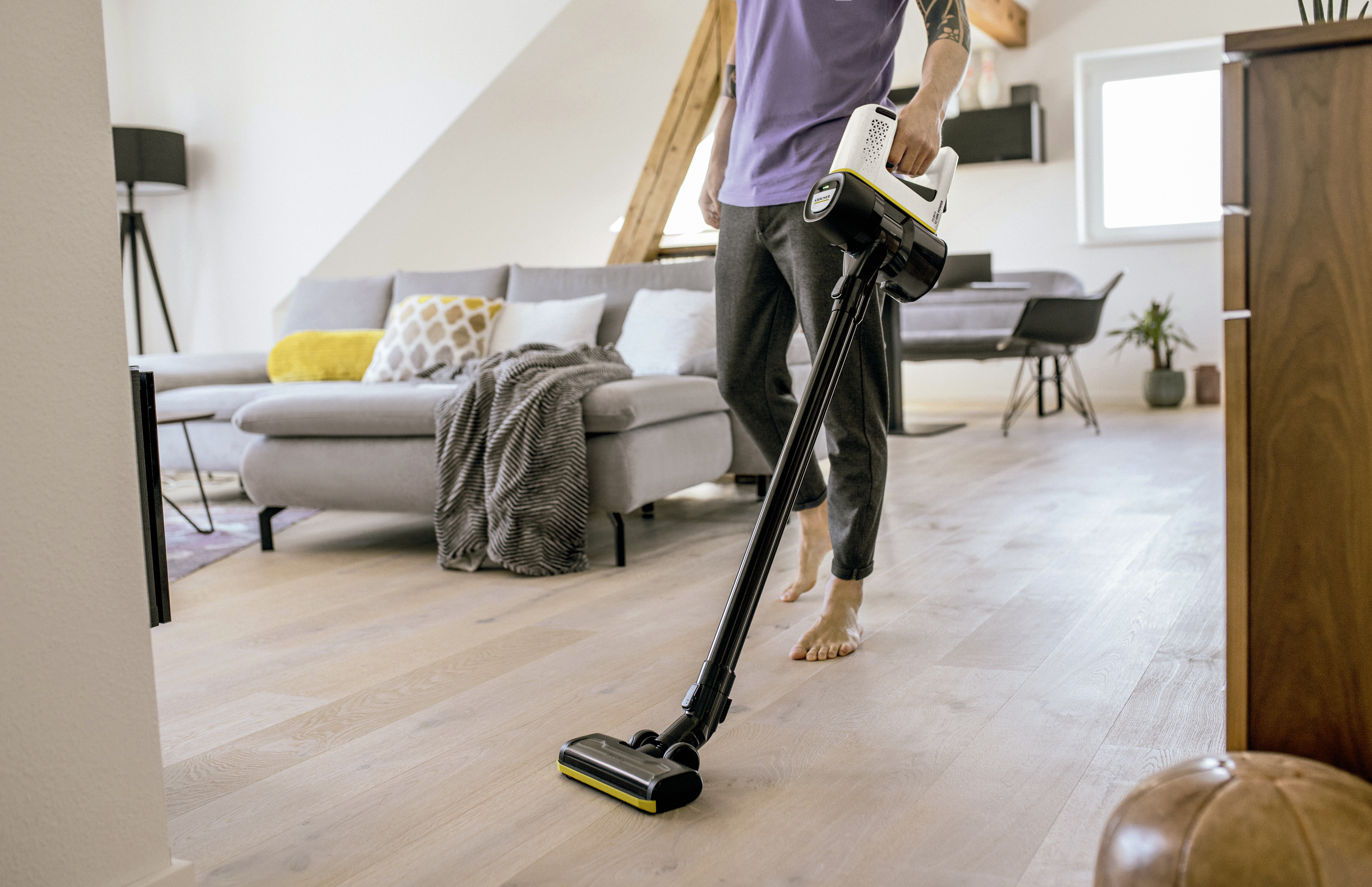 Person vacuuming a wood floor. Sofa, rug, and furniture visible in the background.