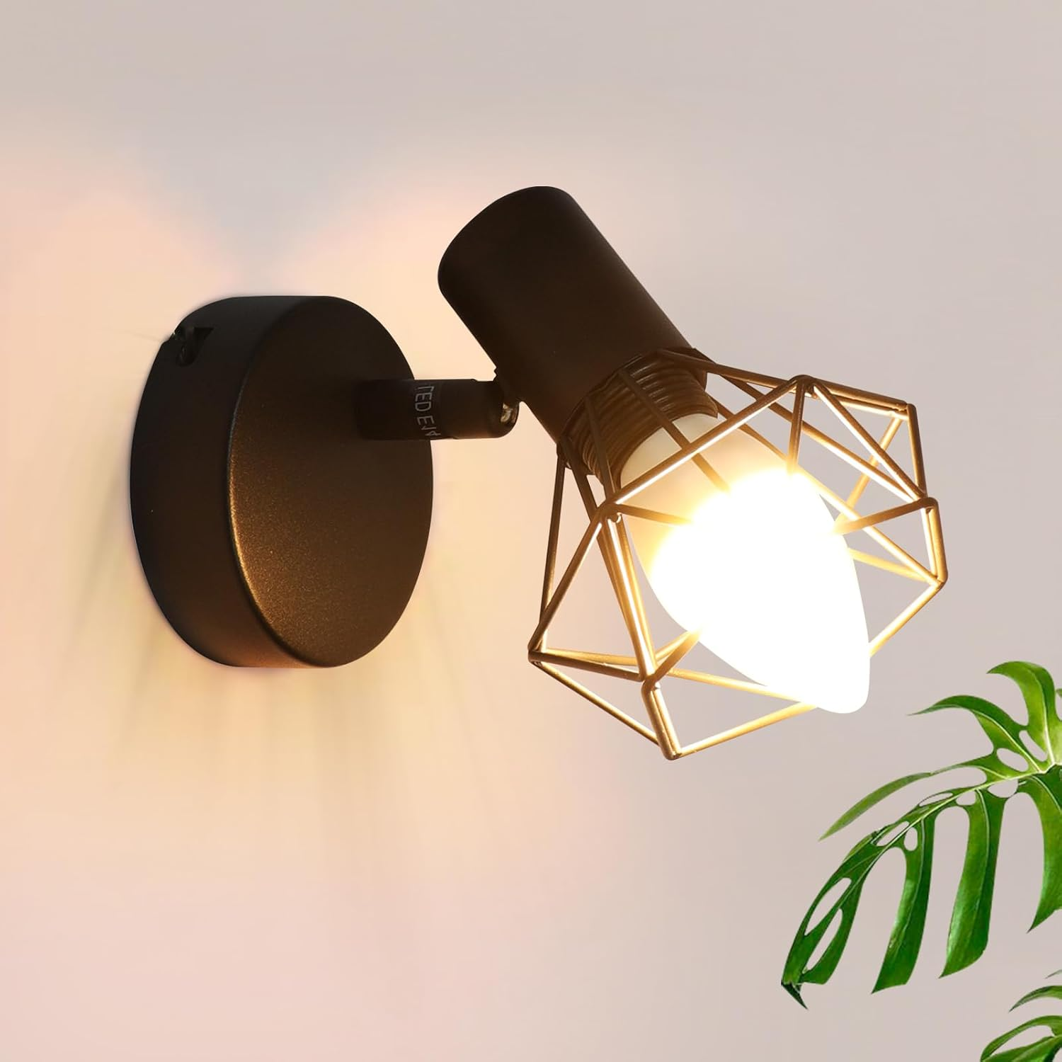 Faretto nero a parete con gabbia geometrica attorno alla lampadina. Montato su muro bianco.