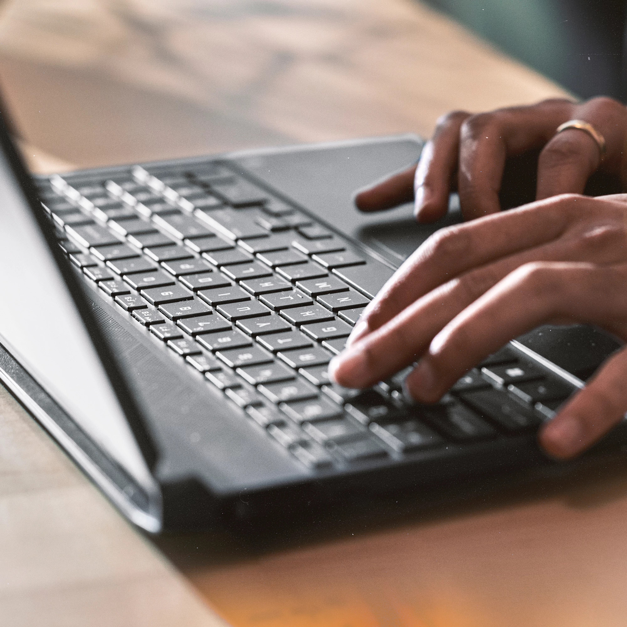 Dłonie piszące na klawiaturze laptopa. Widoczny złoty pierścionek. Jasny drewniany stół.