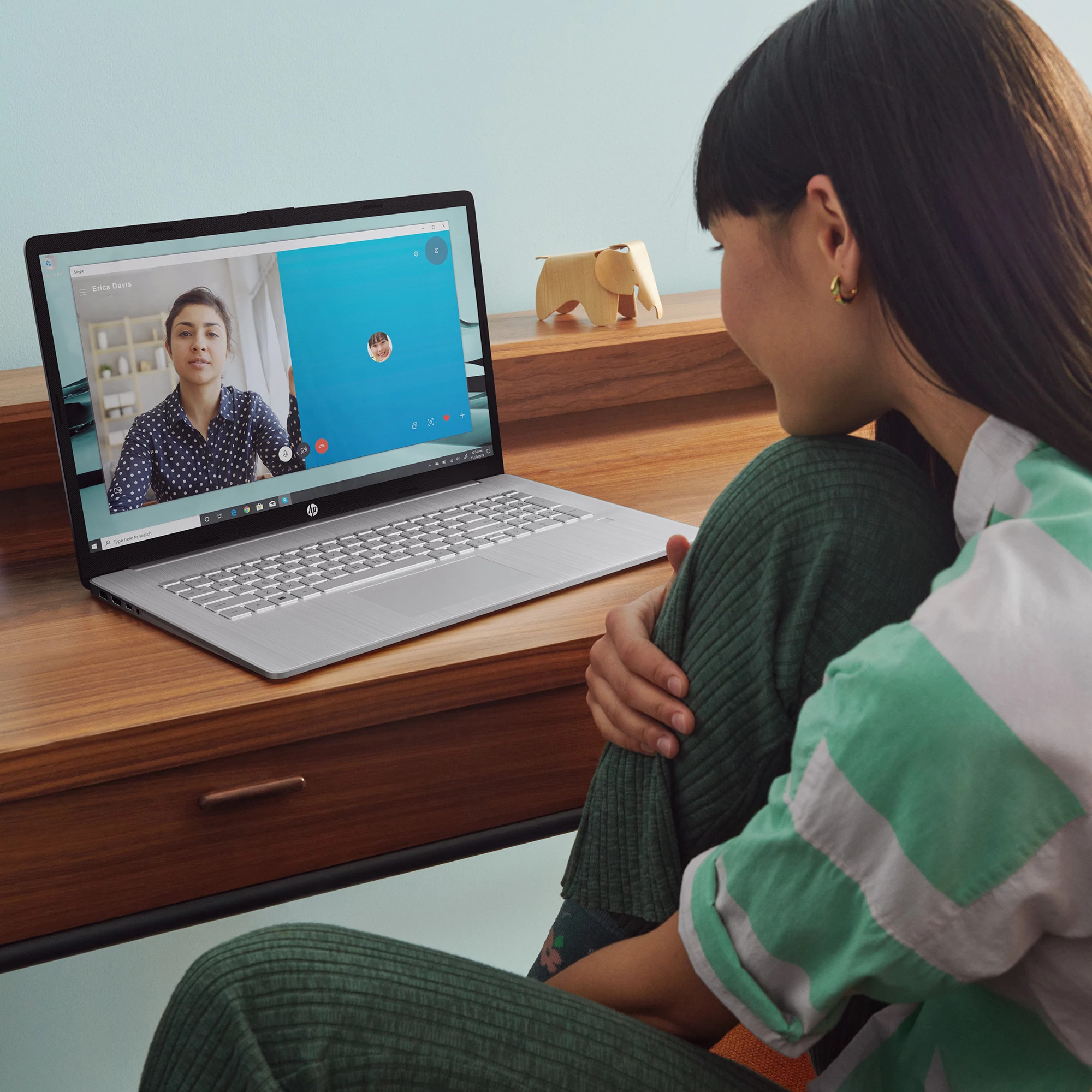 Frau im Videoanruf, Laptop auf Holzschreibtisch. Holz-Elefant und türkisfarbene Wand im Hintergrund.