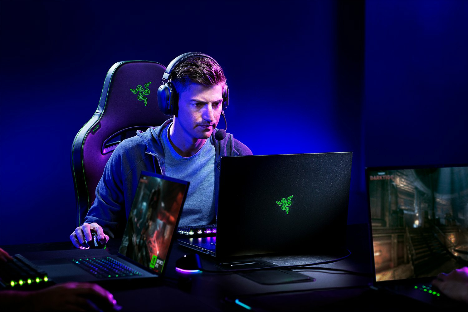 Gamer mit Headset und Laptop, zwei weitere Bildschirme auf dem Schreibtisch, dunkelblau und violett.