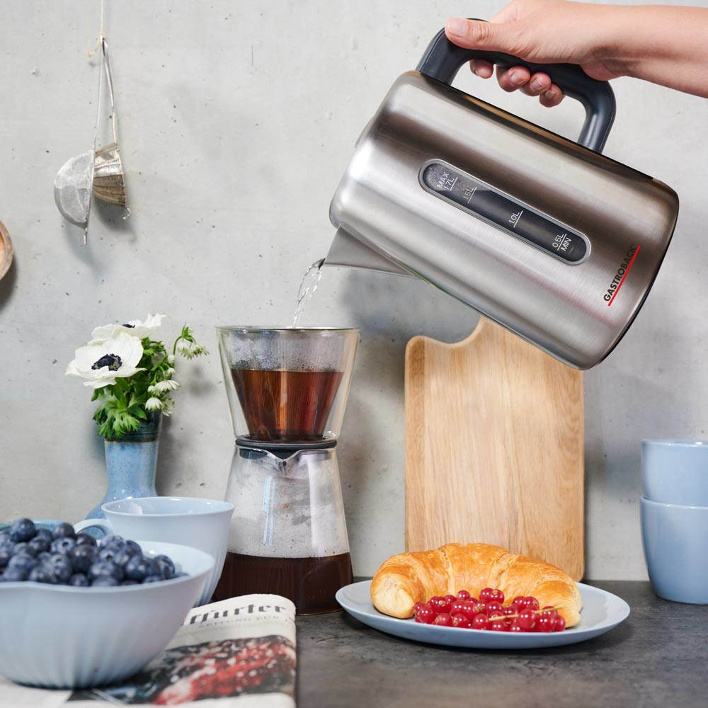 Osoba nalewa wodę ze srebrnego czajnika elektrycznego do szklanego ekspresu do kawy, z produktami śniadaniowymi.