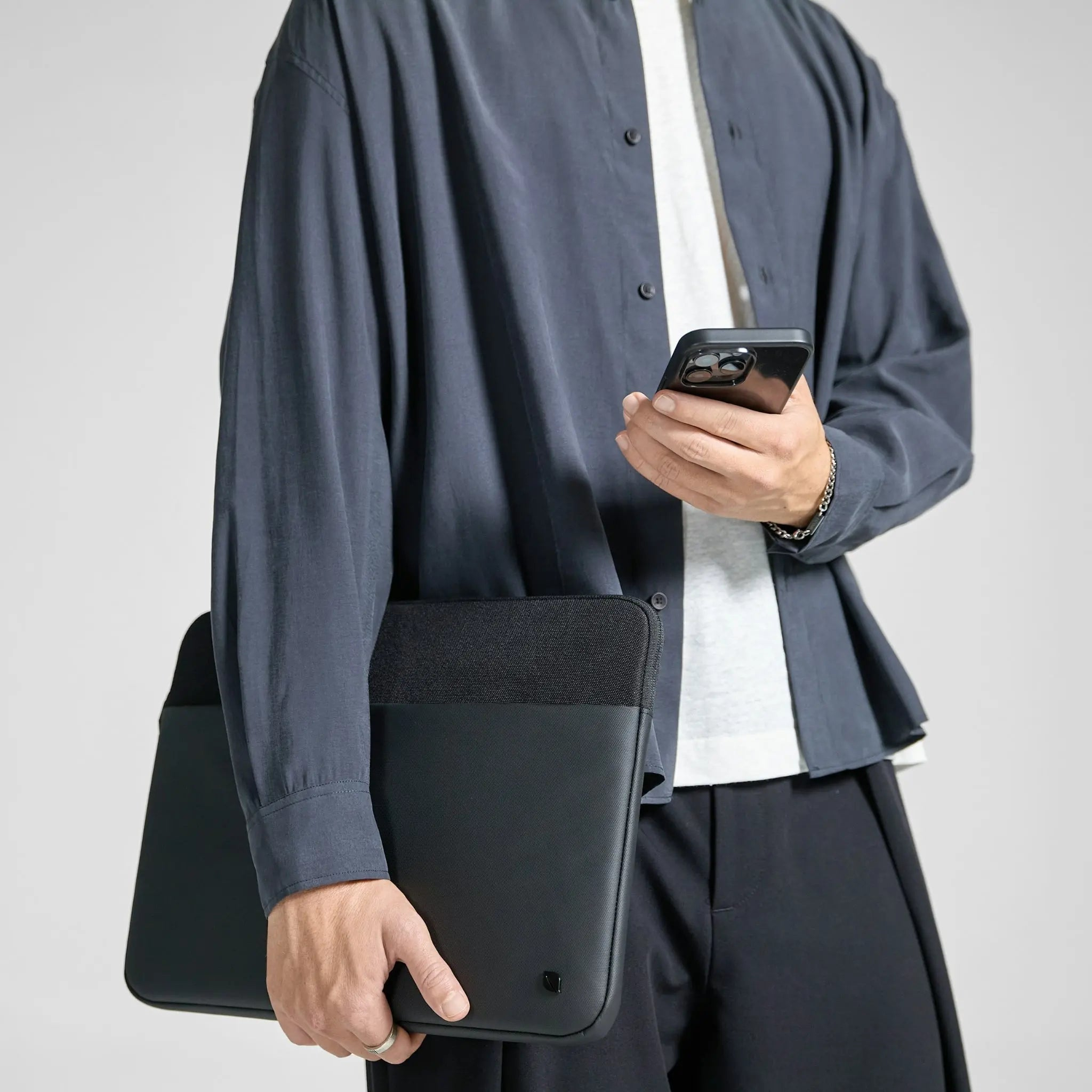 Un uomo tiene una custodia per laptop nera e un telefono. Indossa una camicia blu scuro e pantaloni neri.
