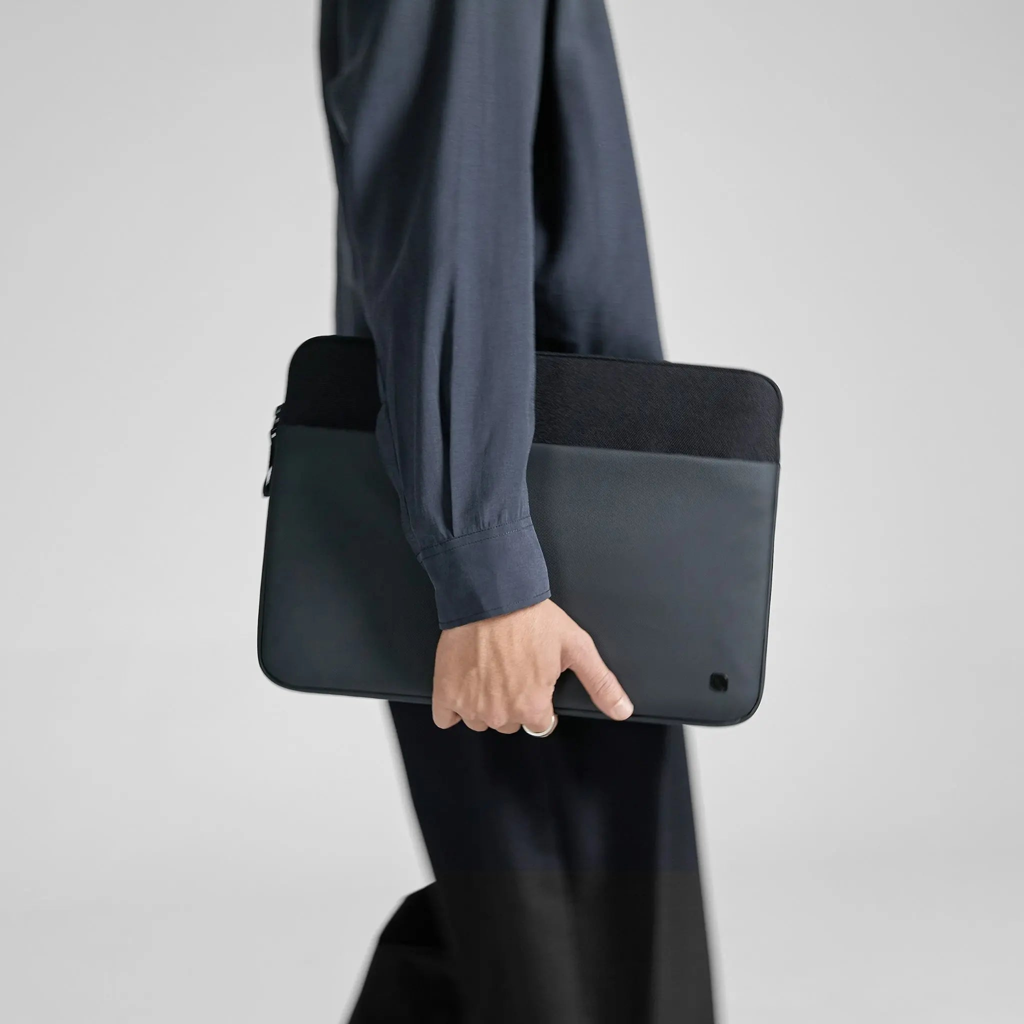 Una persona tiene in mano una custodia nera per laptop. Indossa una camicia e pantaloni blu scuro. Lo sfondo è bianco.