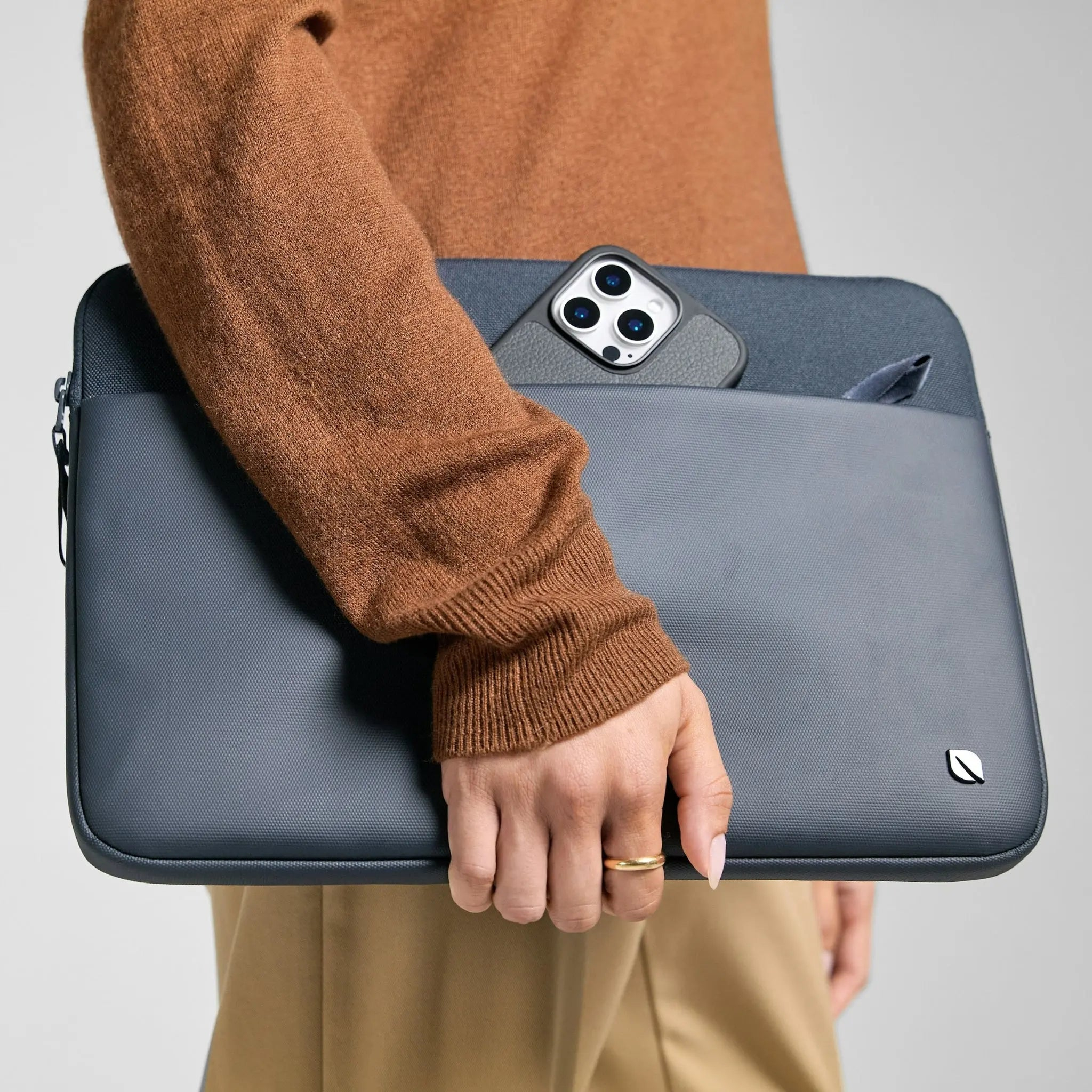 Persona che tiene una custodia grigia per laptop con telefono in tasca. Maglione marrone.