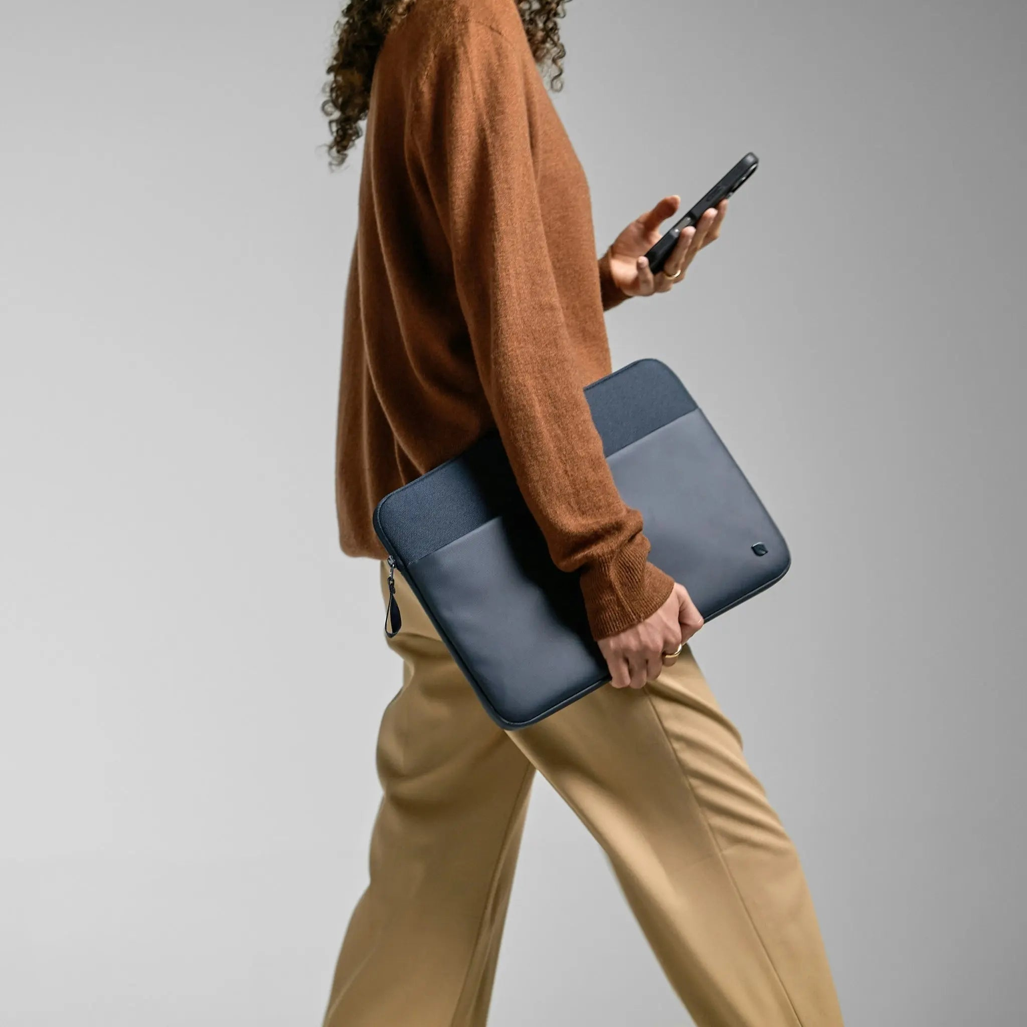 Persona che cammina, tenendo una custodia per laptop blu navy e un telefono, con un maglione marrone e pantaloni beige.