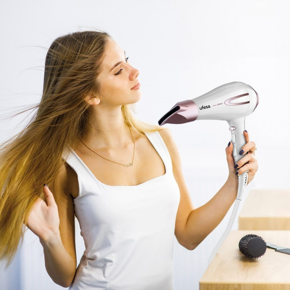 Kobieta używająca białej i różowo-złotej suszarki do włosów, z rozwianymi włosami, w pobliżu drewnianej powierzchni.