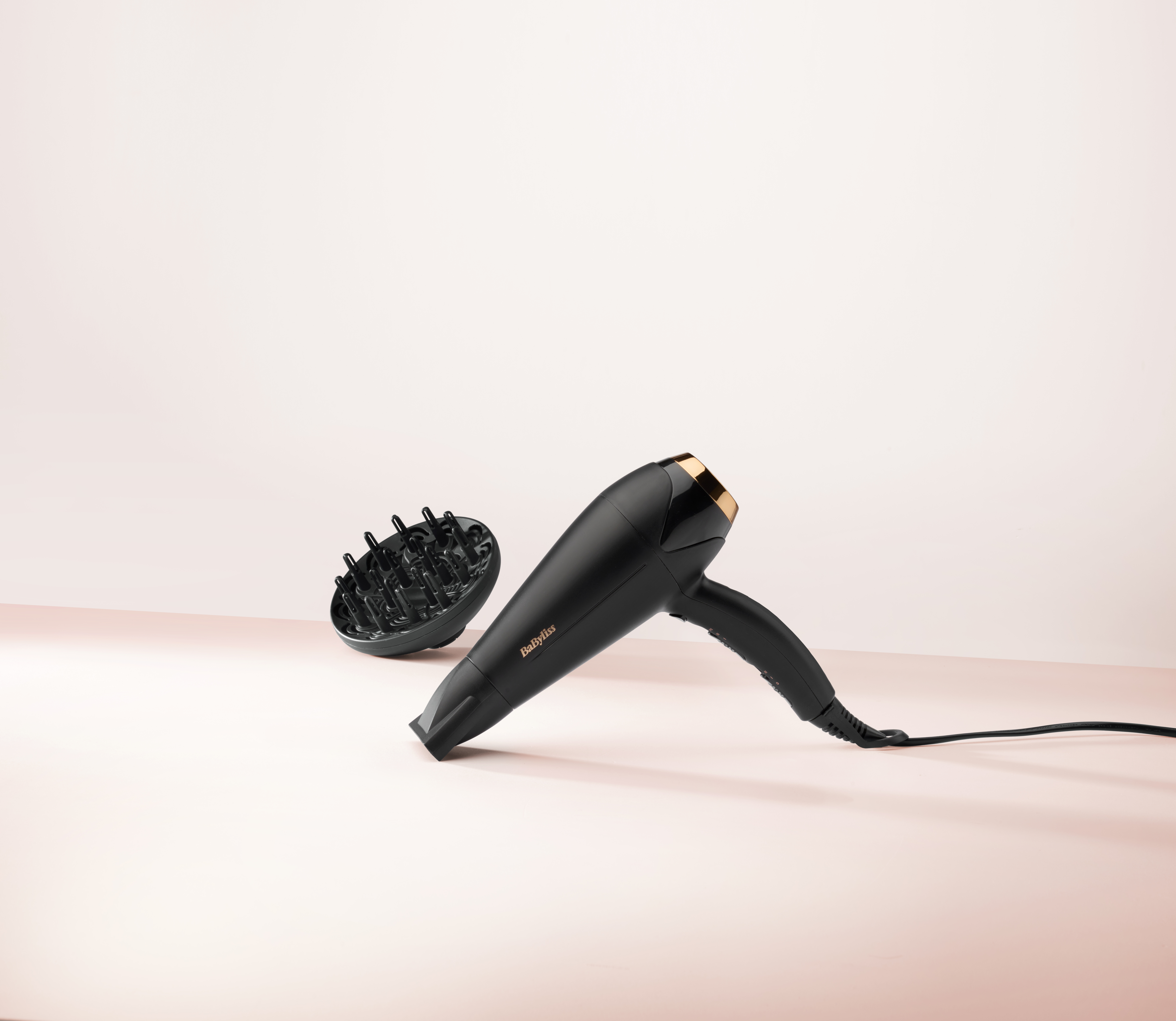 Secador de pelo negro con detalles dorados y difusor sobre superficie rosa.
