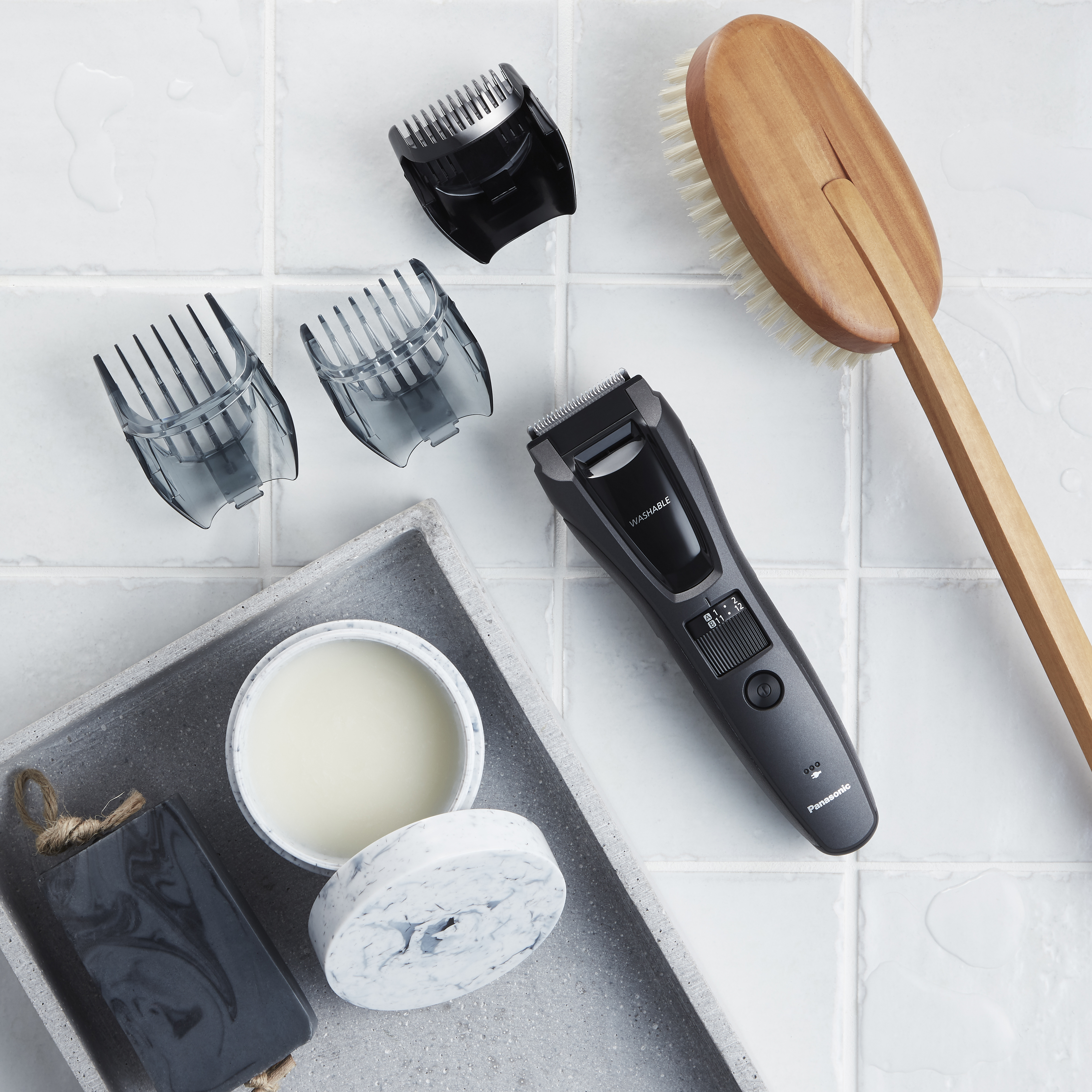 Przybory łazienkowe: trymer, nasadki, szczotka, mydło i krem na białej kafelkowej powierzchni.