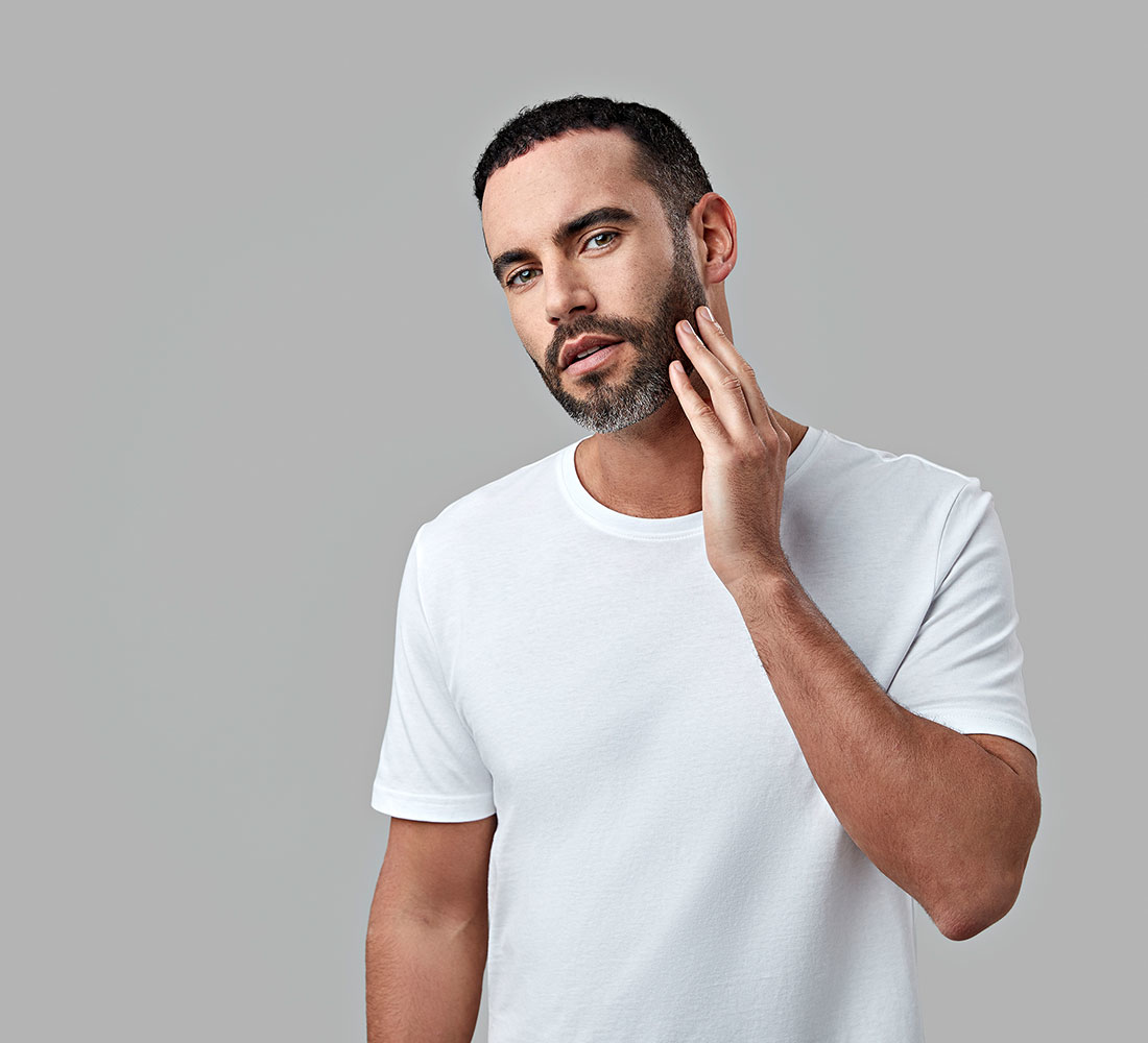 Mann in weißem T-Shirt berührt Bart. Grauer Hintergrund.