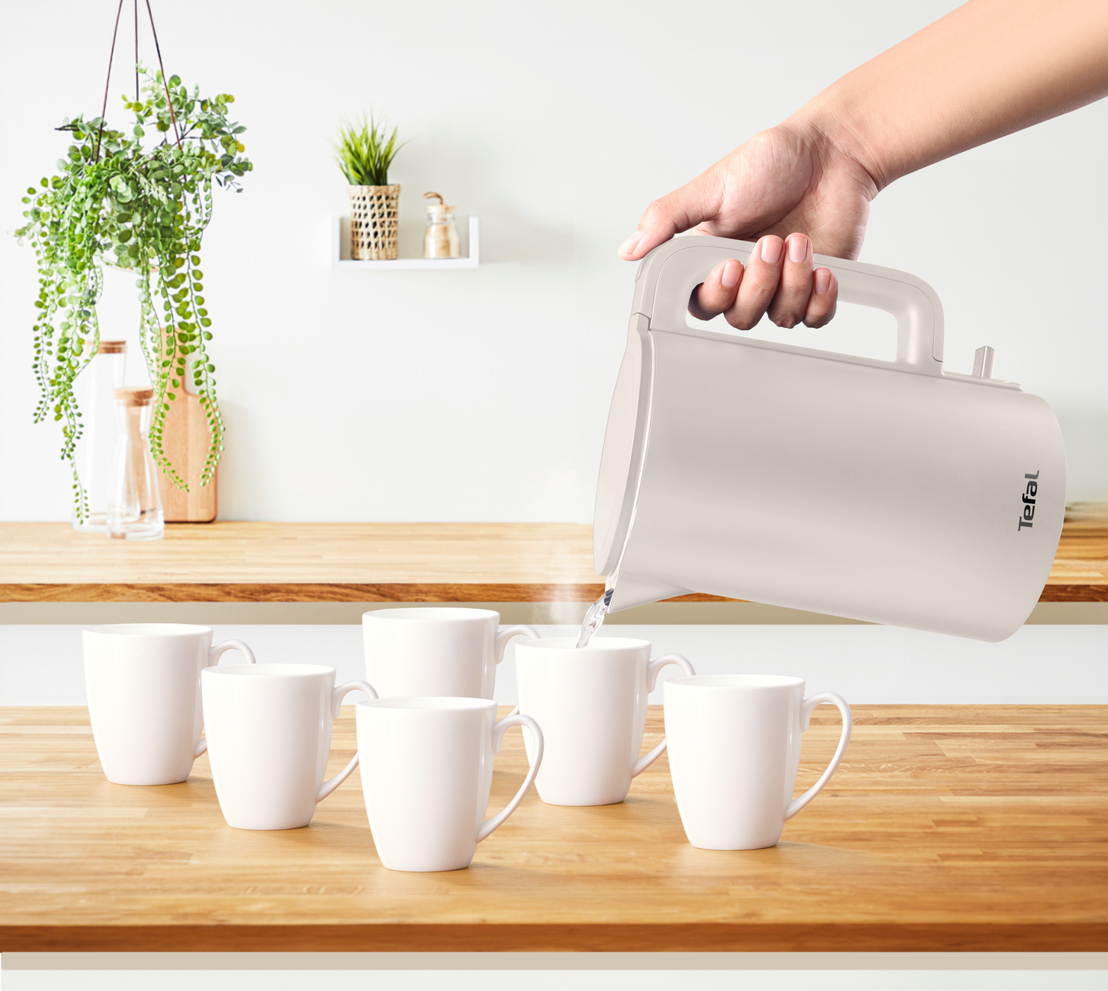 A hand pouring water from a white kettle into white mugs on a wooden table.