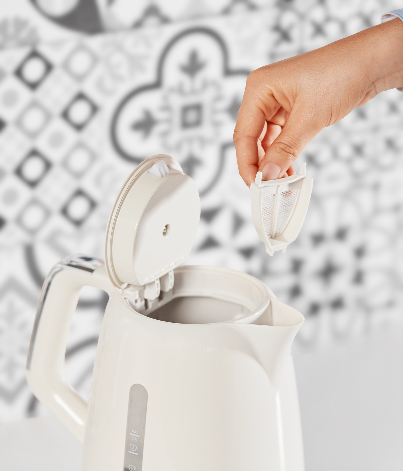 Mano quitando un filtro de té de una tetera blanca. La tetera está abierta. El fondo tiene un patrón.