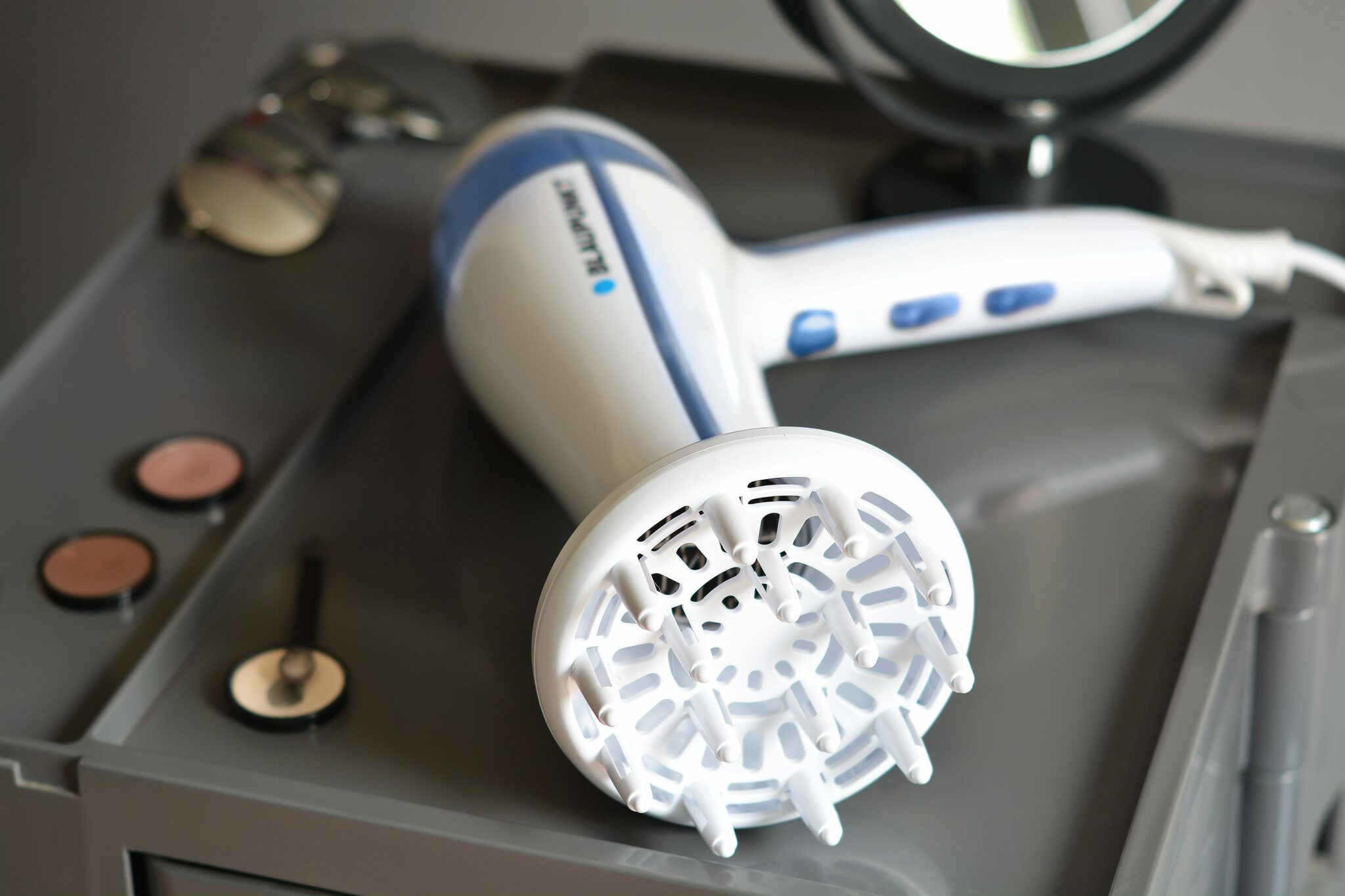 Asciugacapelli bianco con pulsanti blu e diffusore su un carrello grigio. Sono presenti articoli per il trucco.