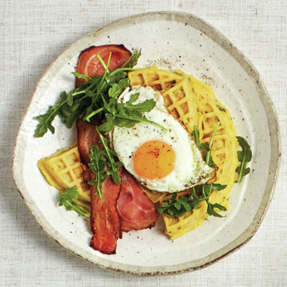 Gofry z jajkiem, boczkiem i rukolą na białym talerzu. Jedzenie jest na białej tkaninie.