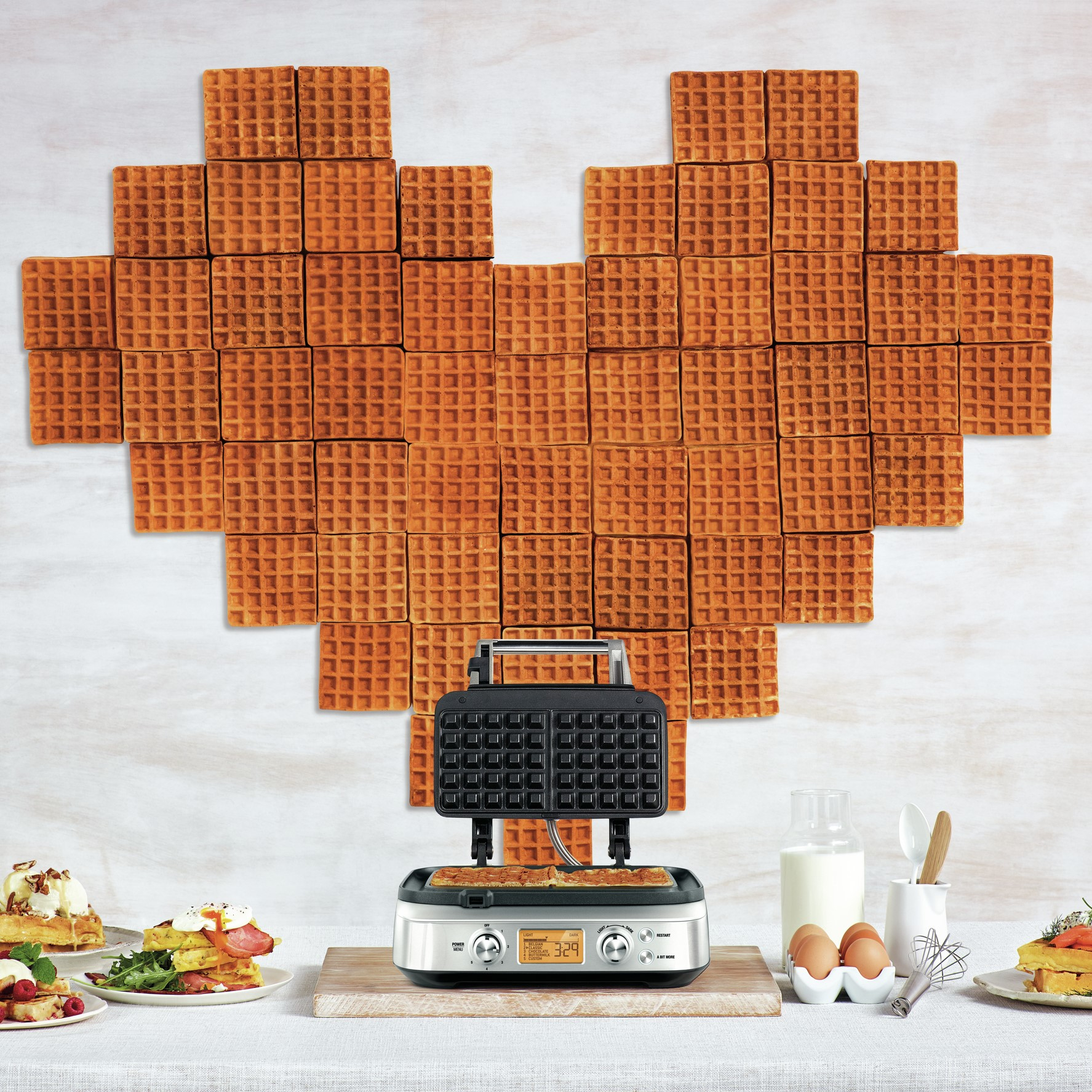 Waffeln in Herzform, Waffeleisen, Eier, Milch und Garnierungen auf dem Tisch.