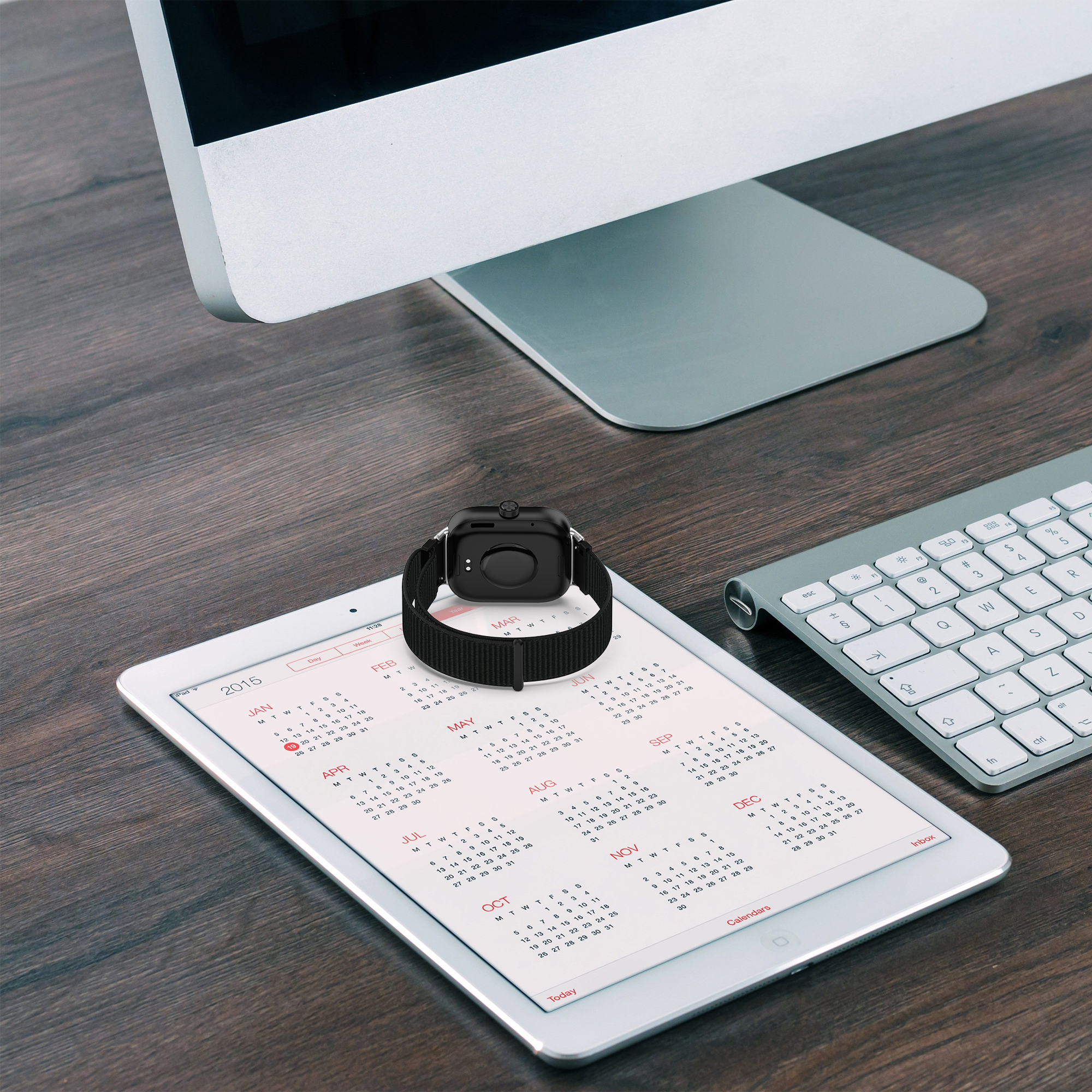 Eine Smartwatch auf einem Kalender-Tablet, in der Nähe eines Monitors und einer Tastatur.