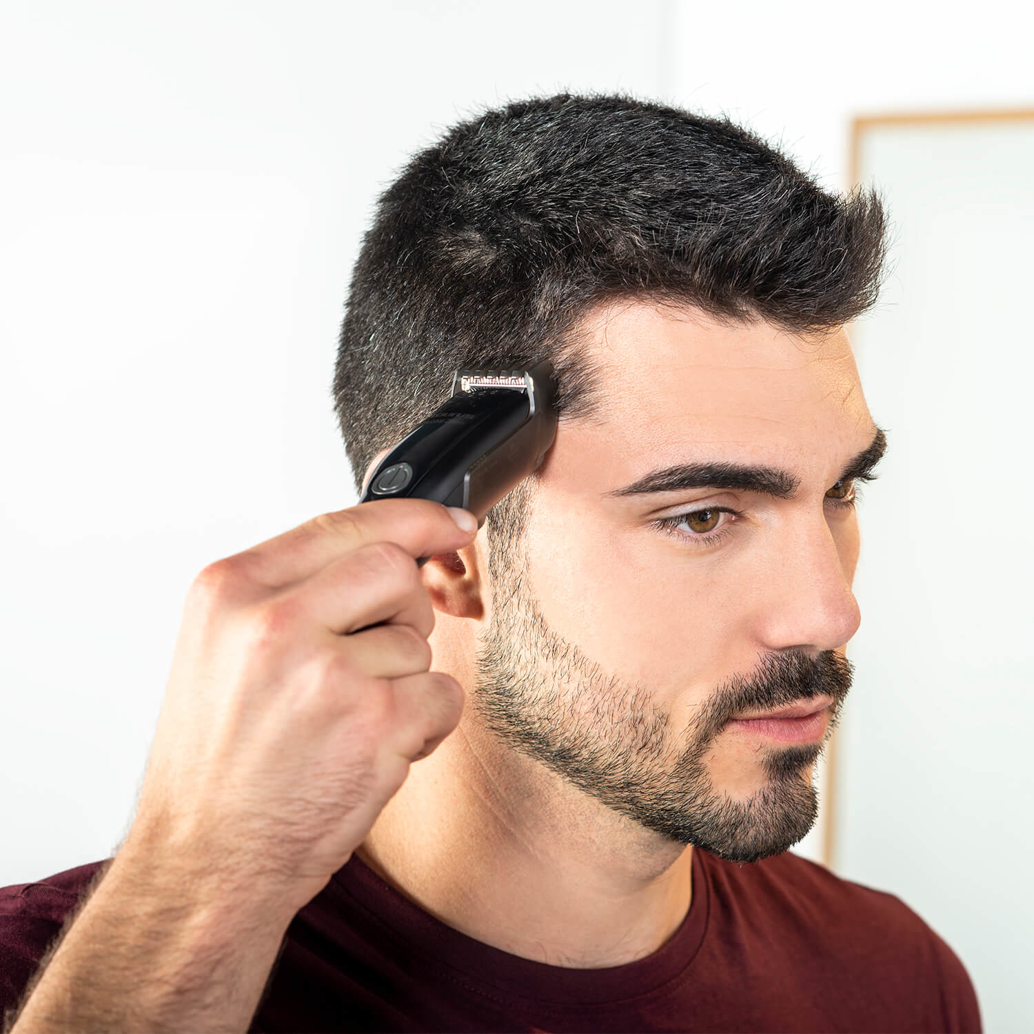 Un uomo che si taglia la barba con un trimmer nero, davanti a un muro bianco.